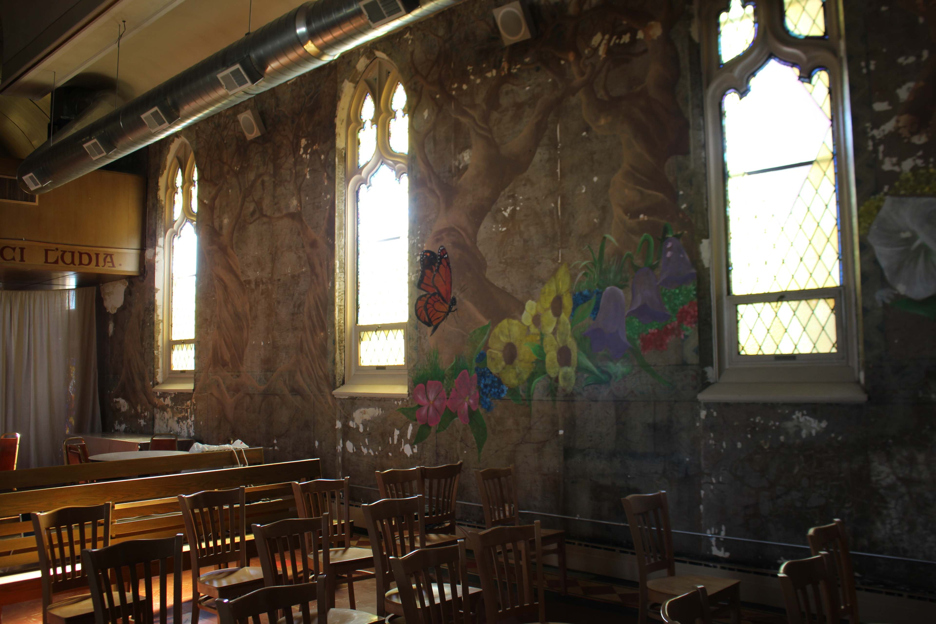 The image shows an interior space with wooden chairs arranged around a wall featuring faded murals and large, arched windows.
