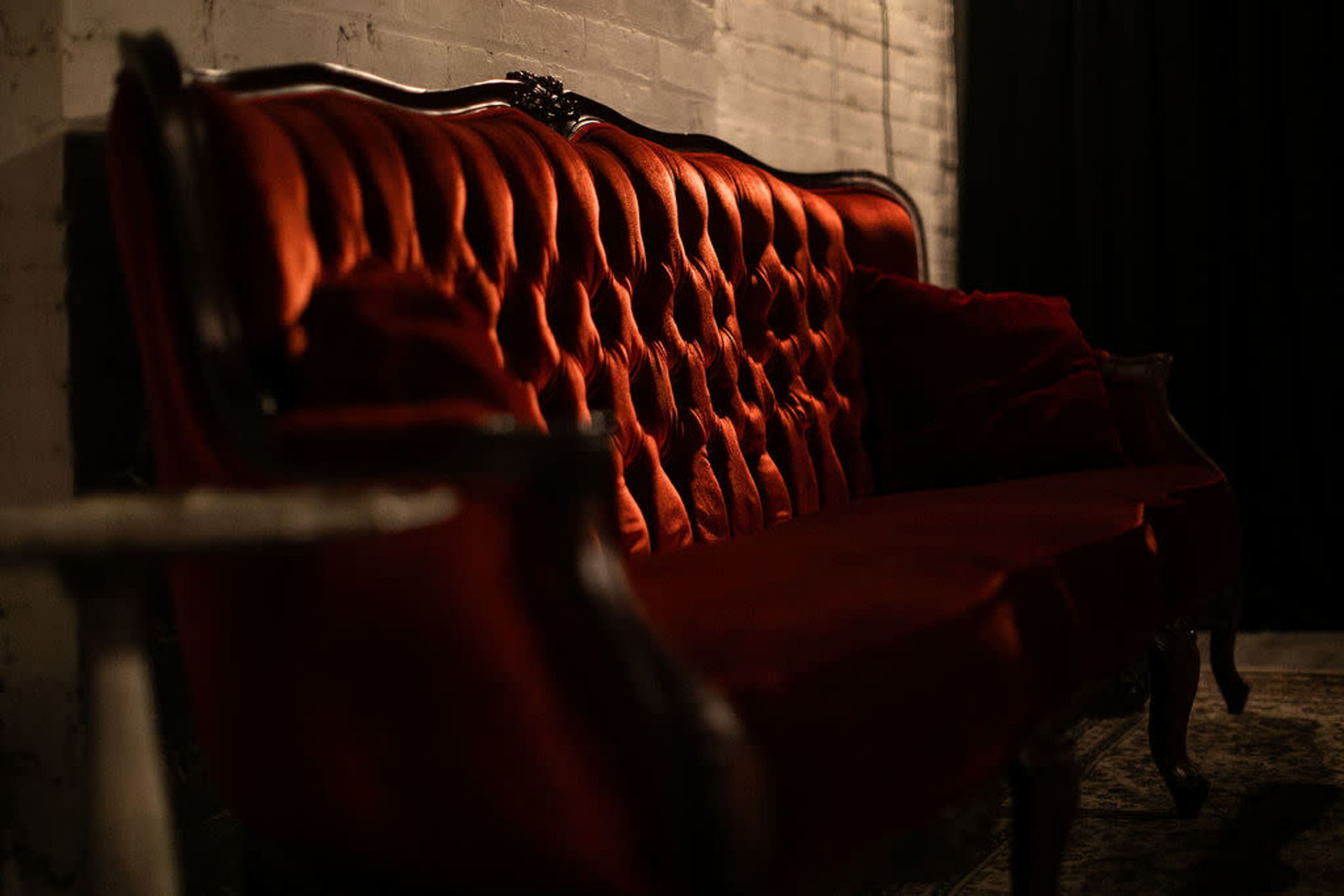A vintage red tufted sofa is positioned against a brick wall in a dimly lit room.