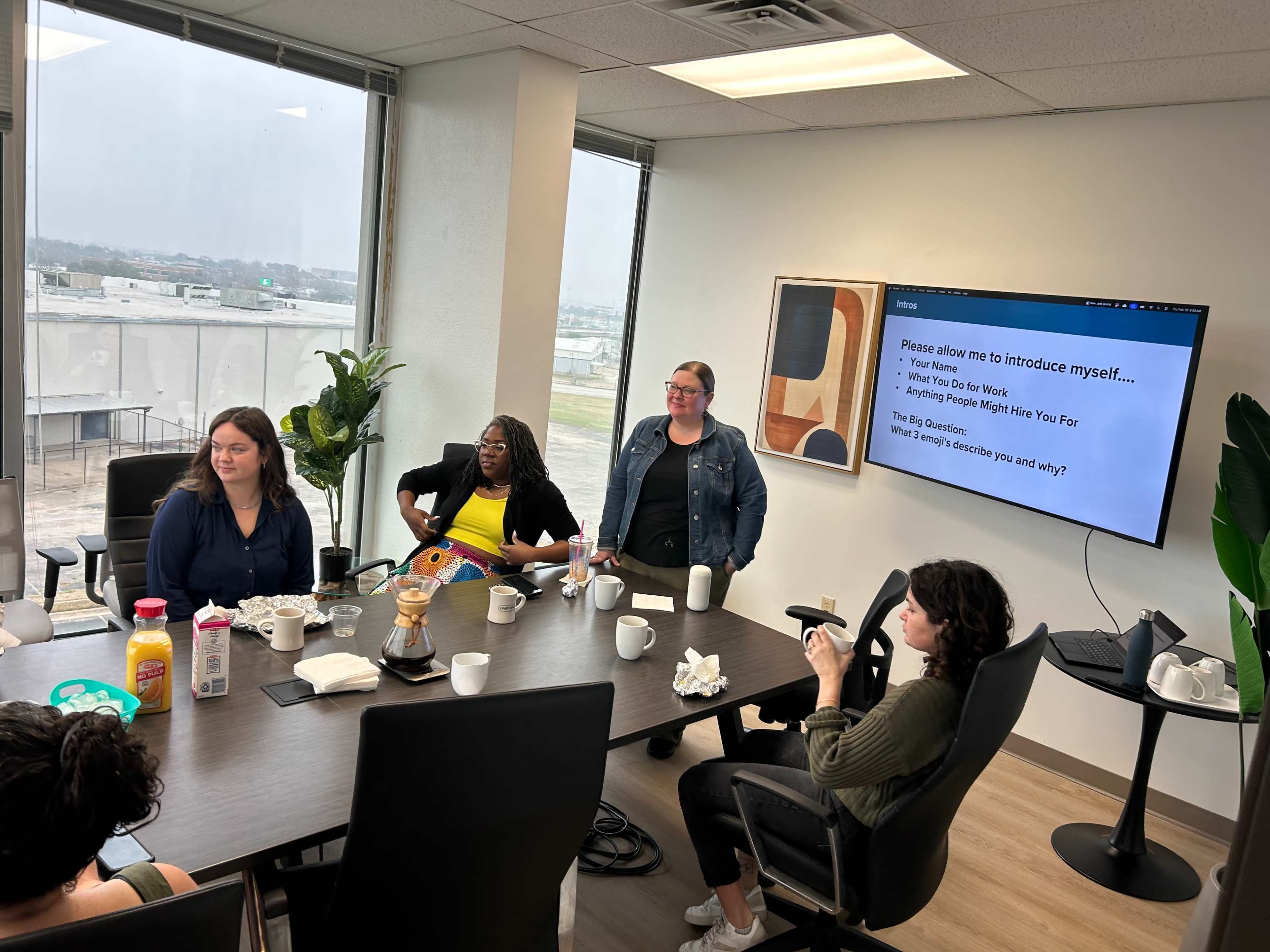 A group of five people are seated around a conference table with breakfast items and a presentation screen displaying an introduction slide.