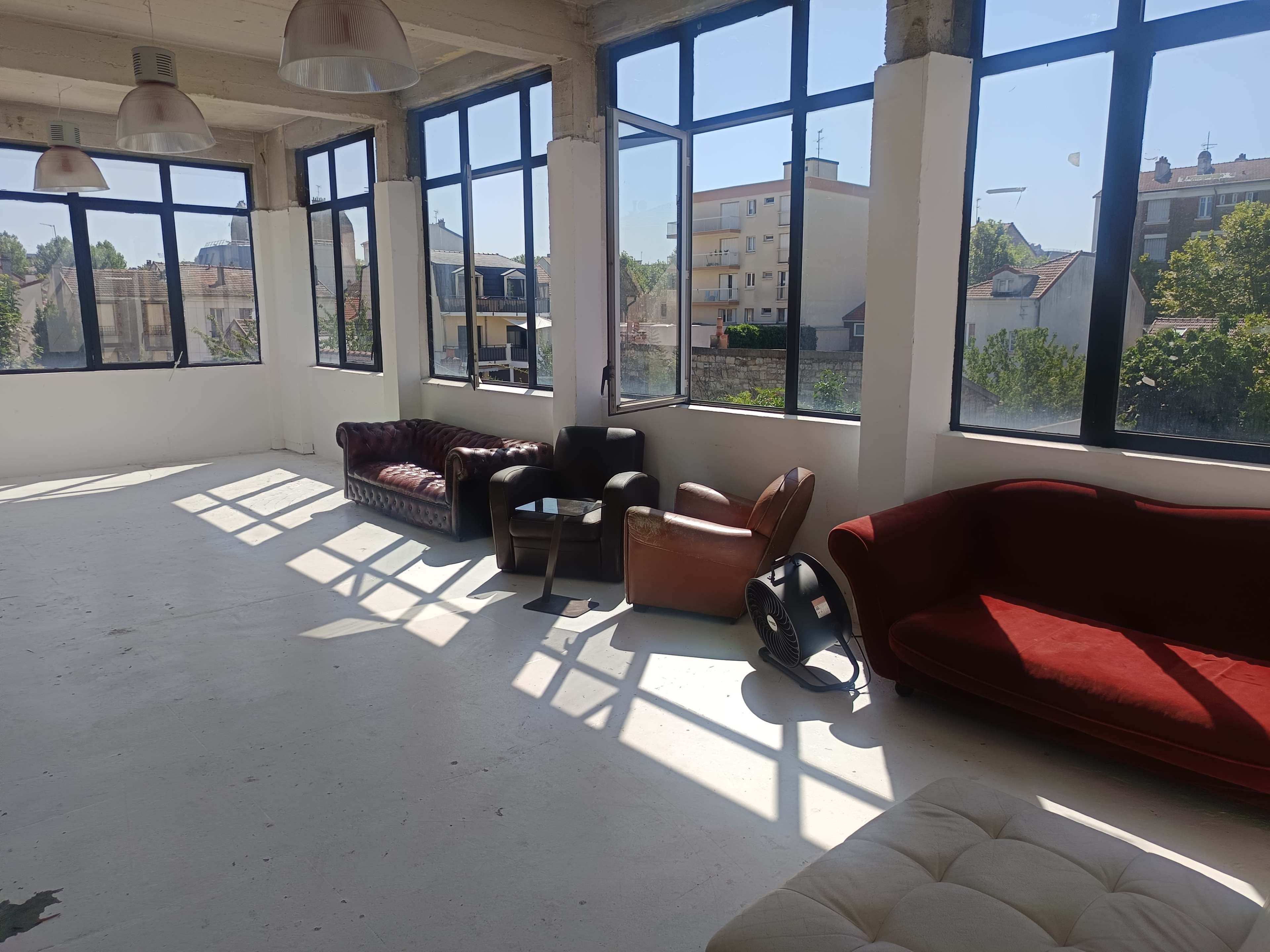 The image shows a bright, sparsely furnished room with large windows, featuring several sofas and a fan.