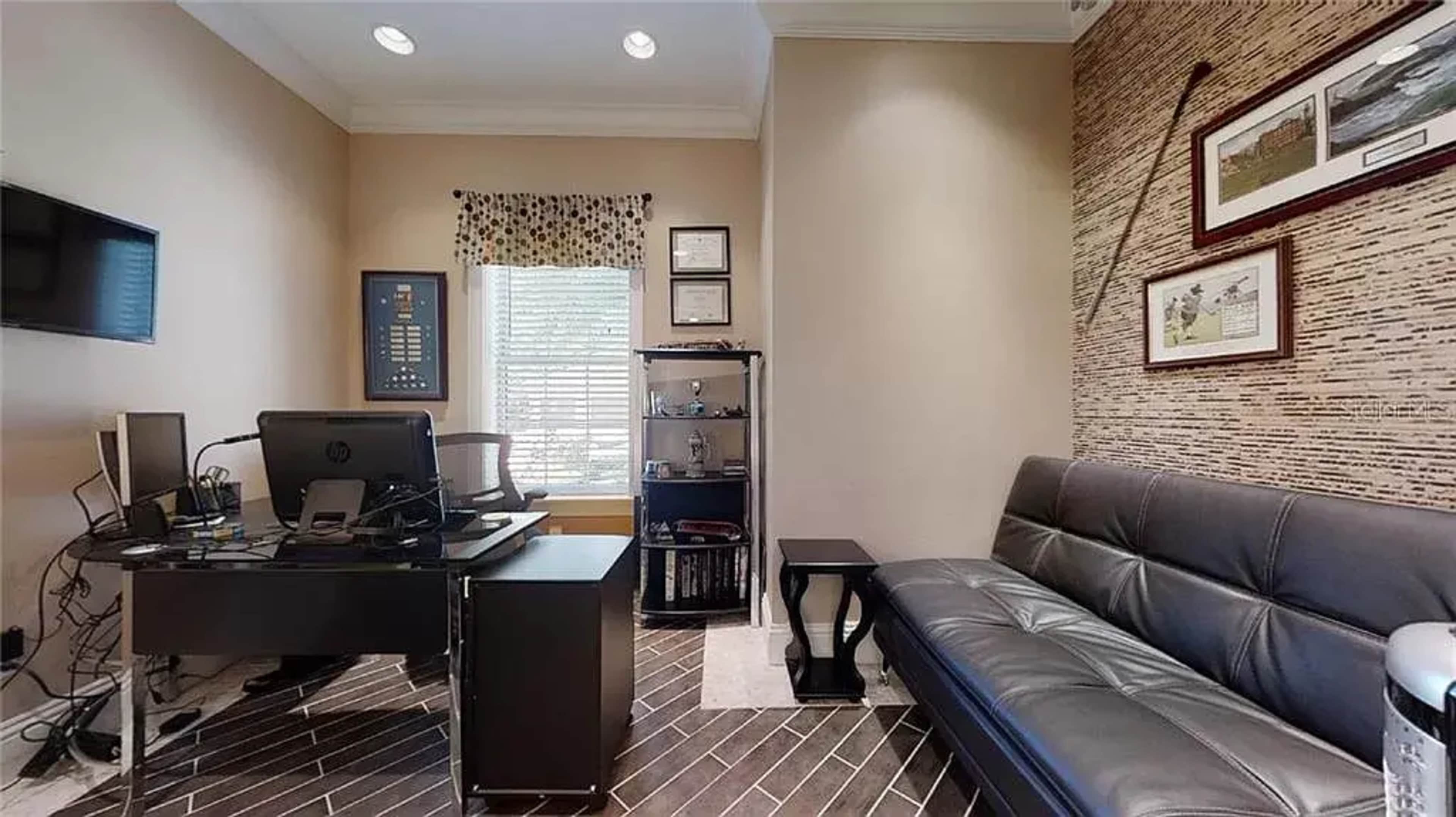 A small office with a desk, chair, and a black couch, featuring a window with a patterned curtain and a wall decorated with framed pictures.