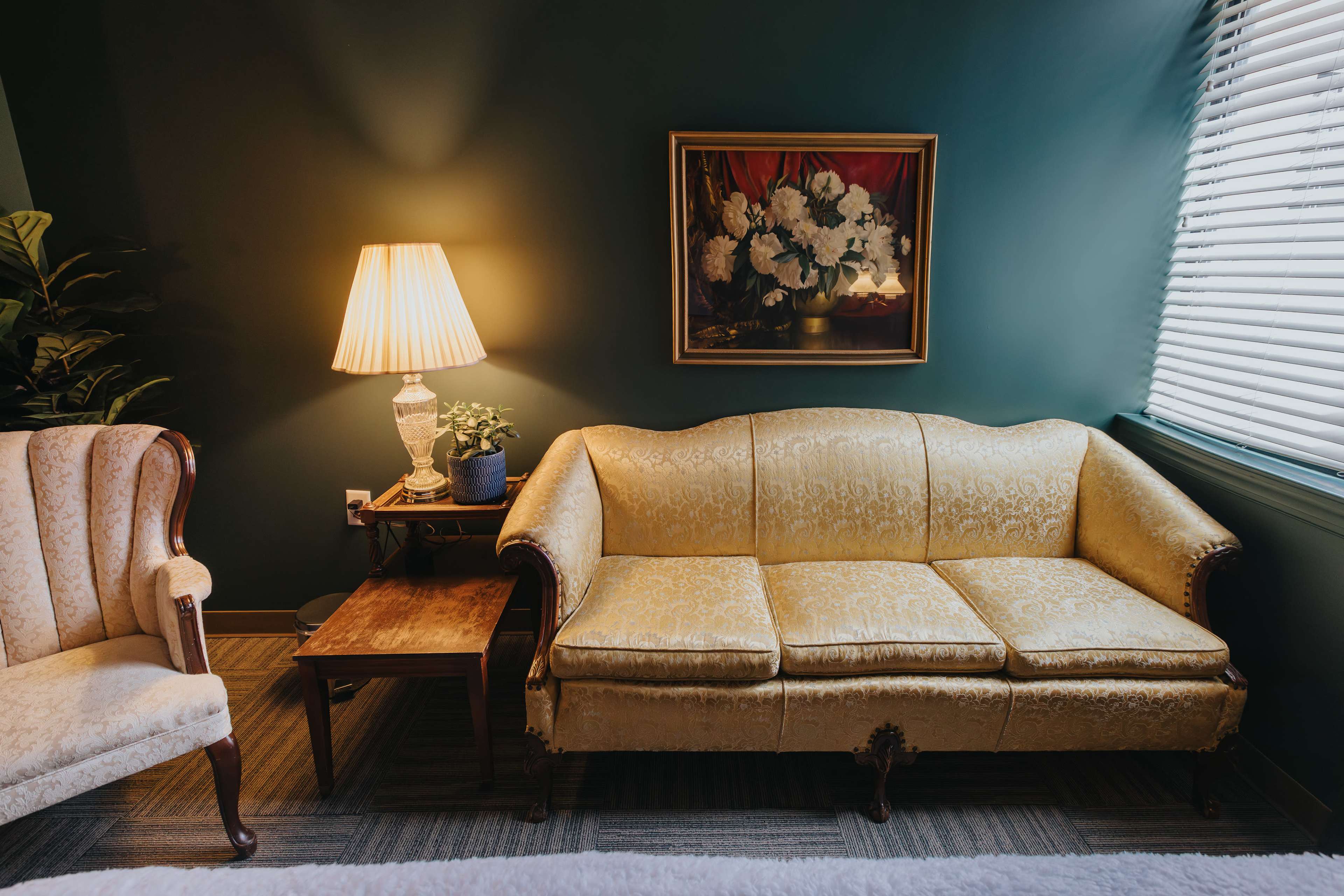 The scene features a vintage yellow sofa beside a lamp and a small table, with a framed painting of flowers on the wall and a window with blinds.