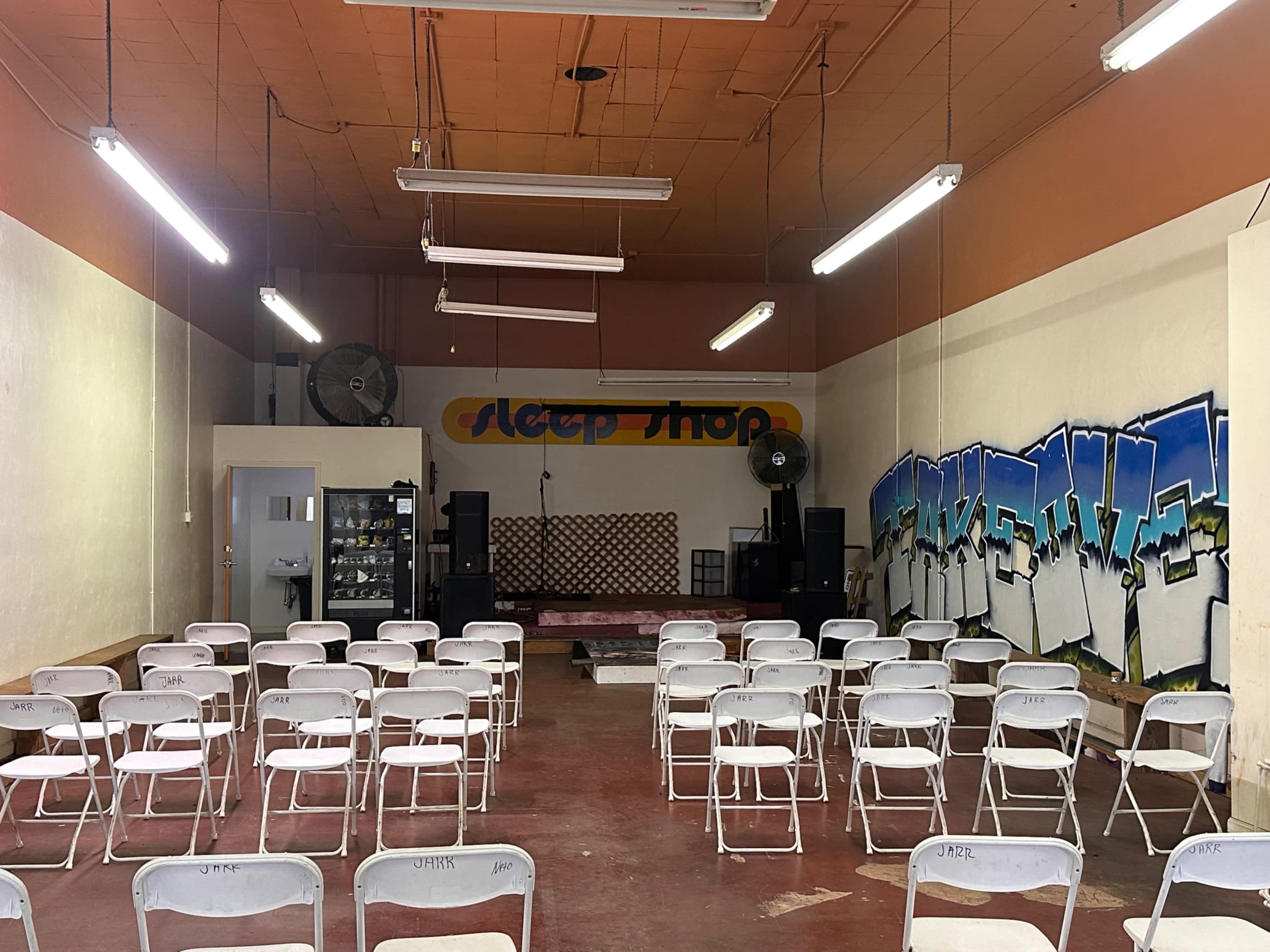 The image shows an empty event space with rows of white chairs facing a small stage, featuring a graffiti wall and a vending machine in the corner.