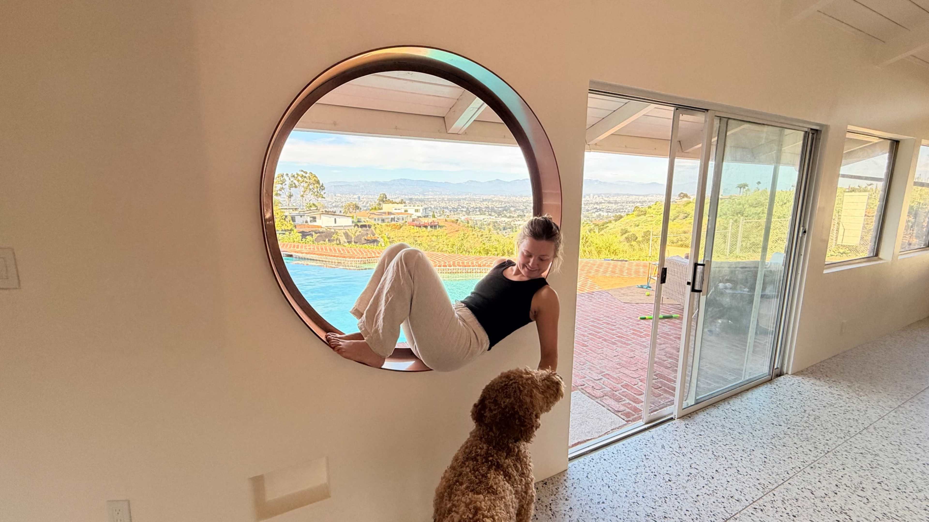 A woman sits in a circular window opening while a dog looks up at her, with a view of a pool and distant mountains in the background.