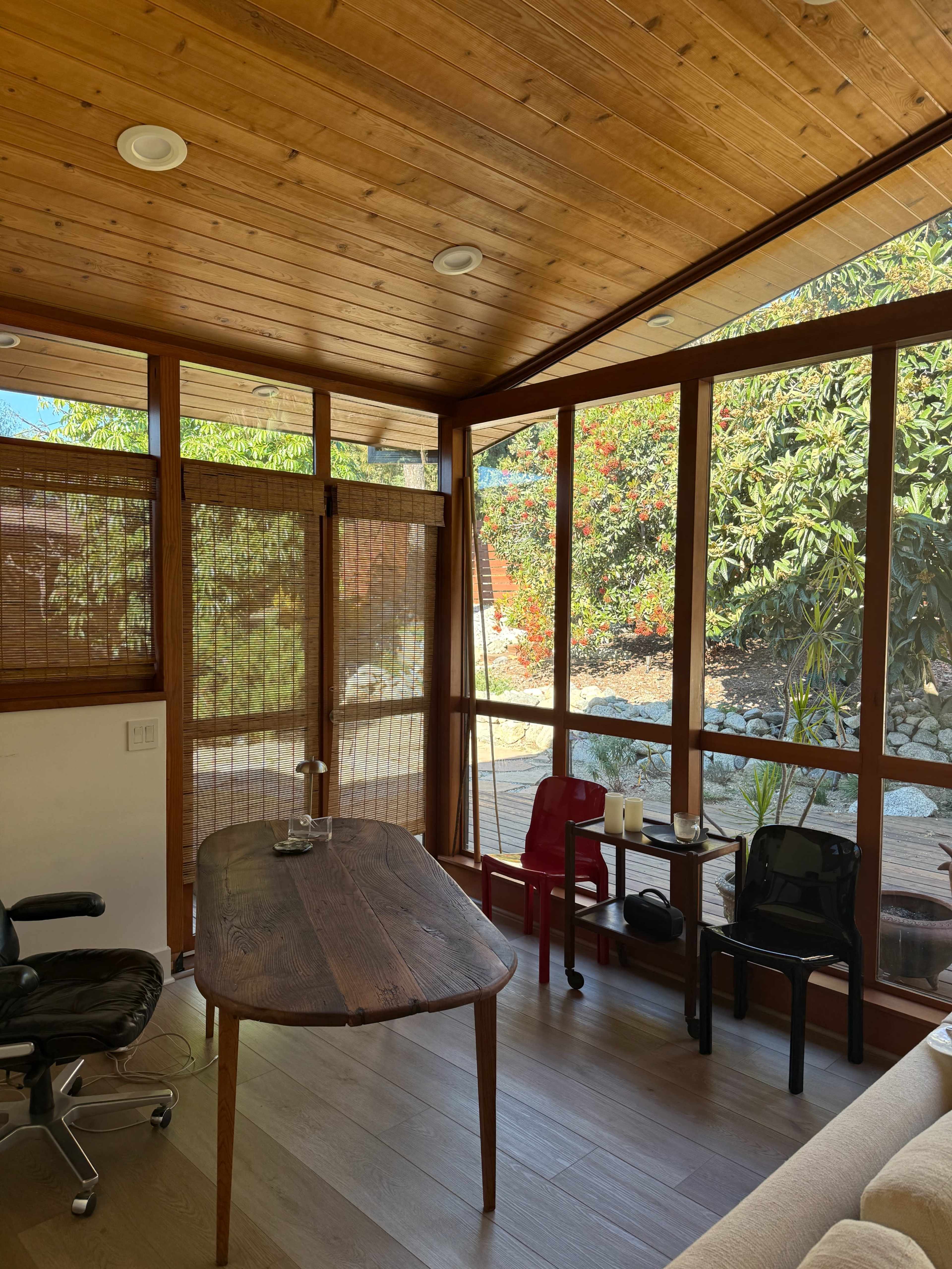The image shows a bright sunroom with wooden paneling, a table, a chair, and large glass windows overlooking a garden.
