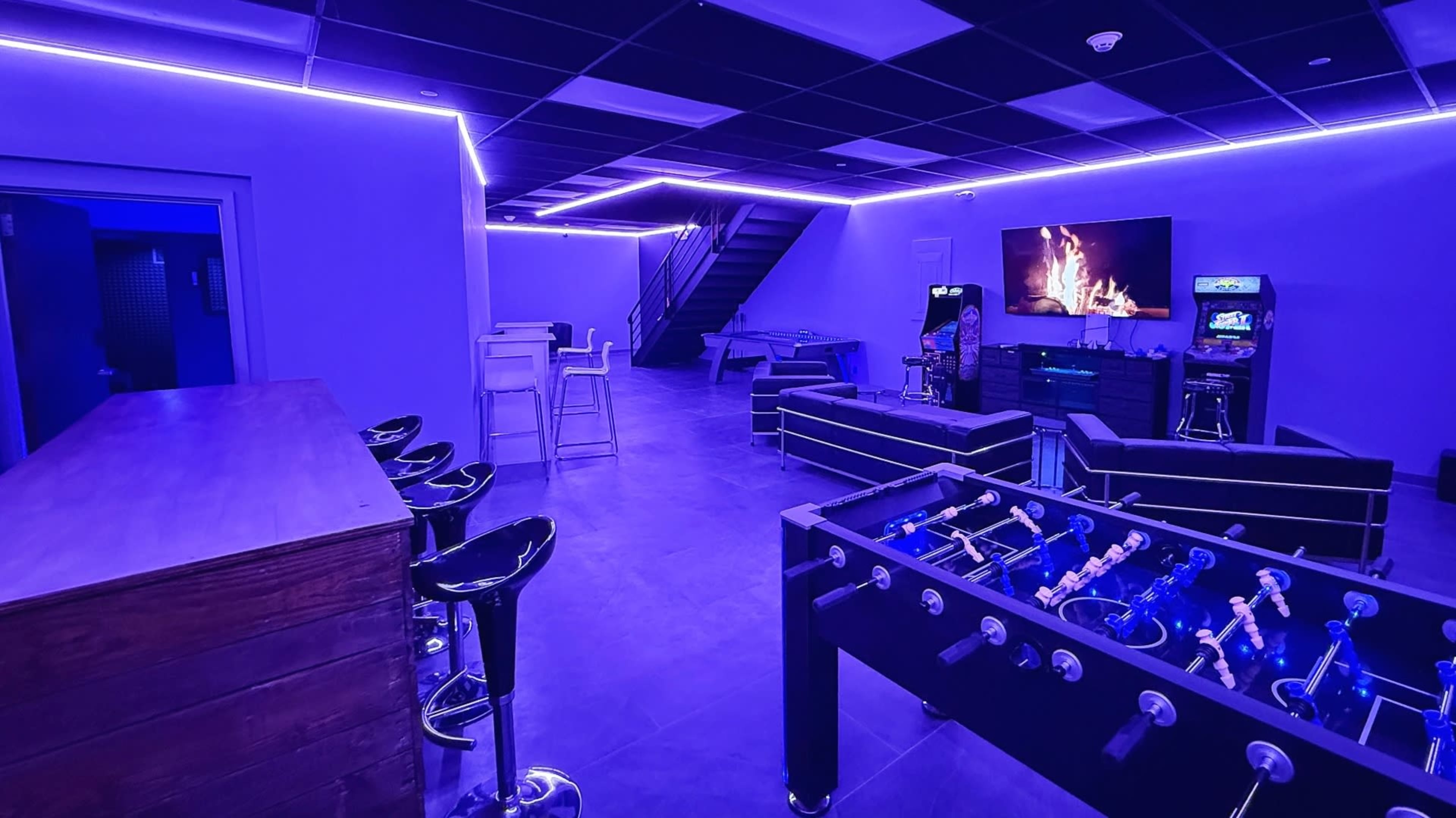 The image shows a modern lounge area with blue LED lighting, featuring a foosball table, seating arrangements, a bar, and a television displaying a fireplace.