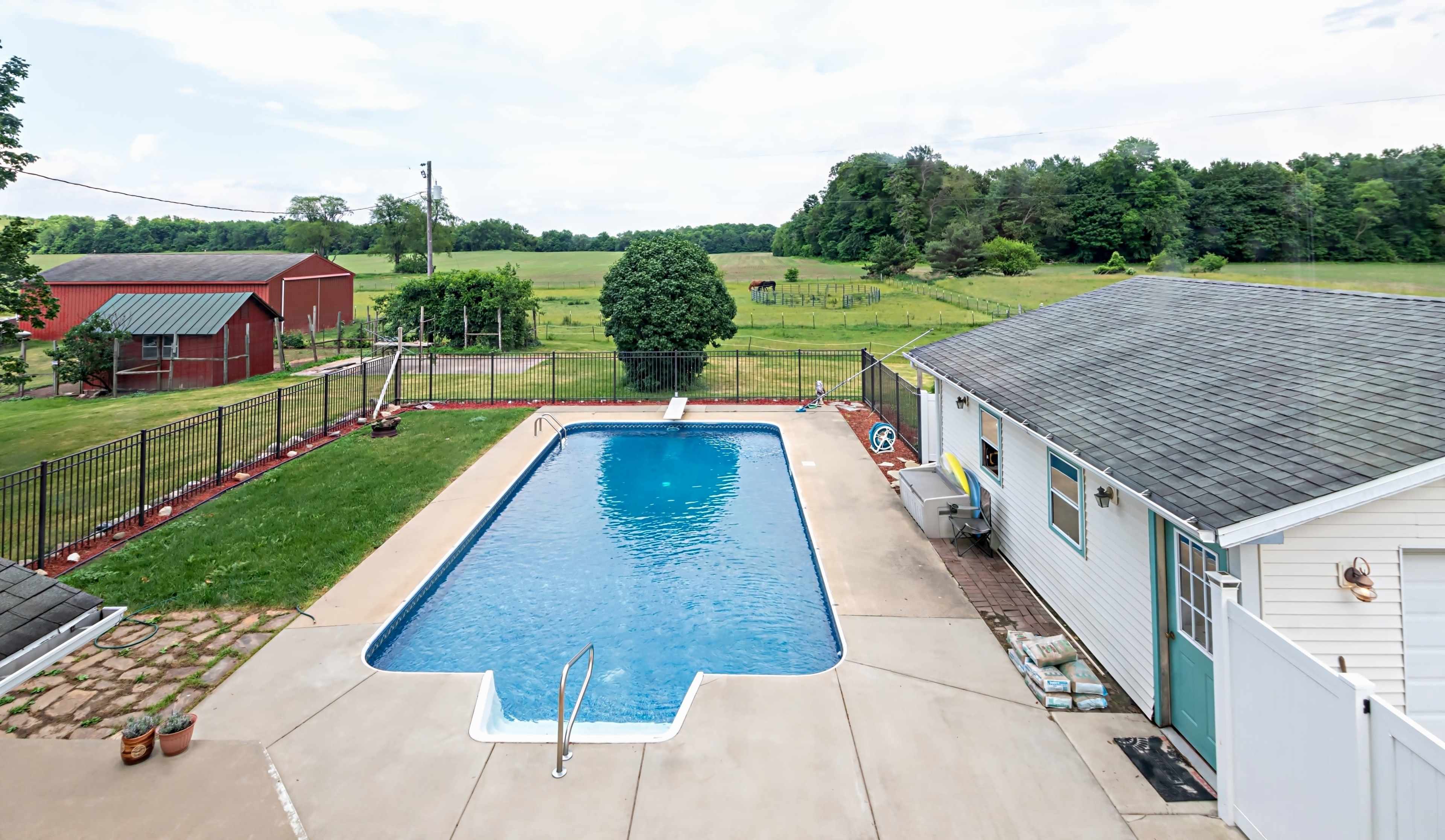 A swimming pool surrounded by a concrete patio, located beside a house and overlooking a green field with a red barn in the background.