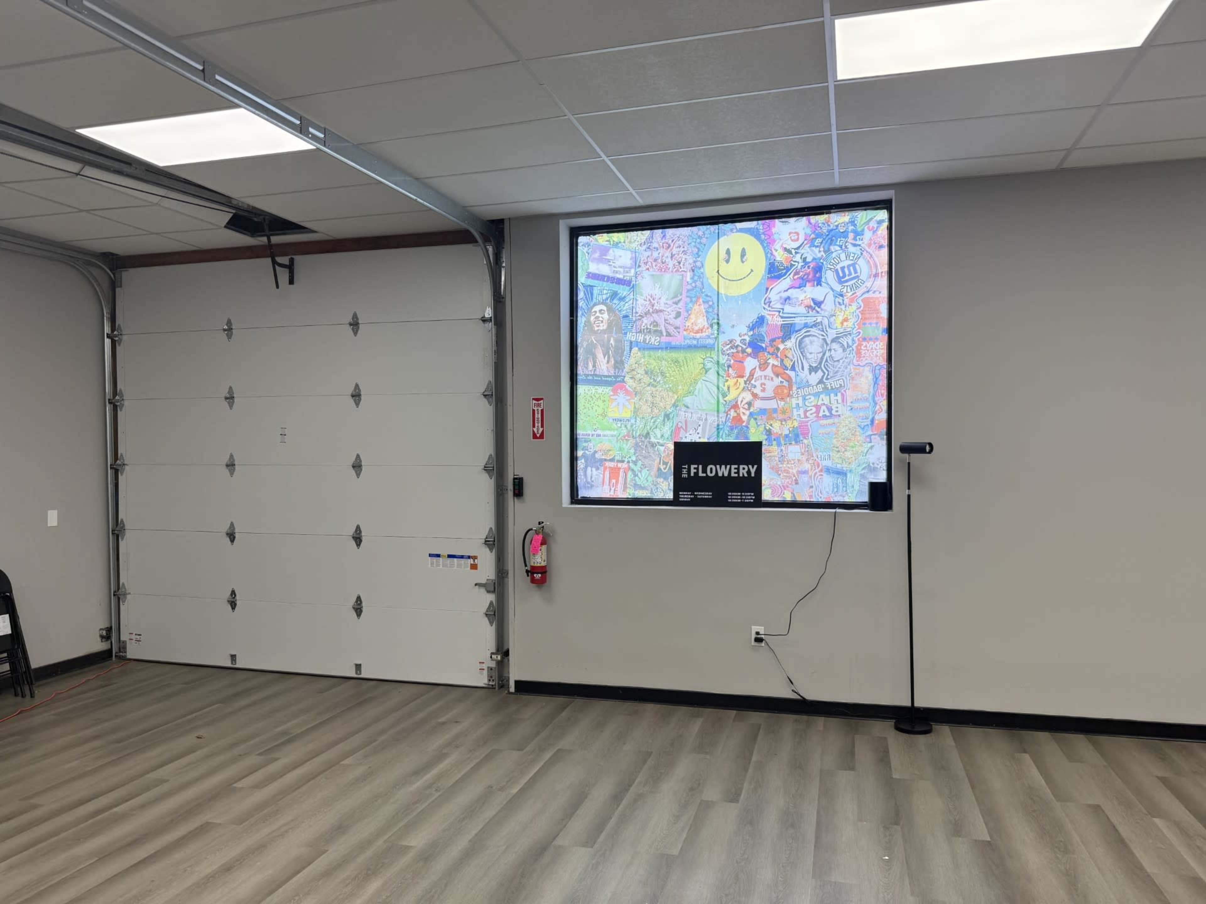 A large screen displaying a colorful collage is mounted on a gray wall in a room with a garage door and a fire extinguisher.