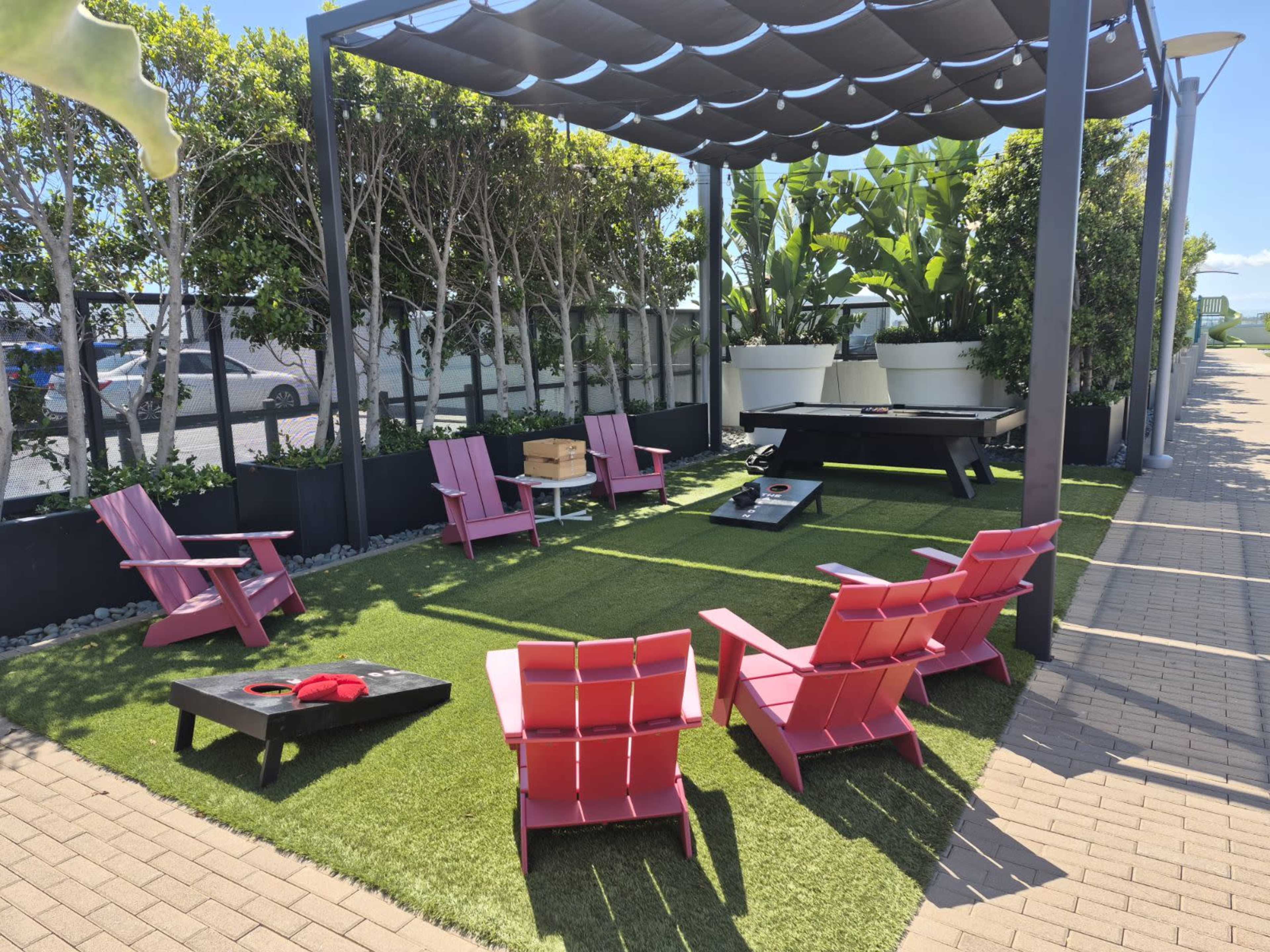 The image shows a lounge area with pink chairs arranged around a central table on a grass-like surface, under a shaded canopy with large potted plants in the background.