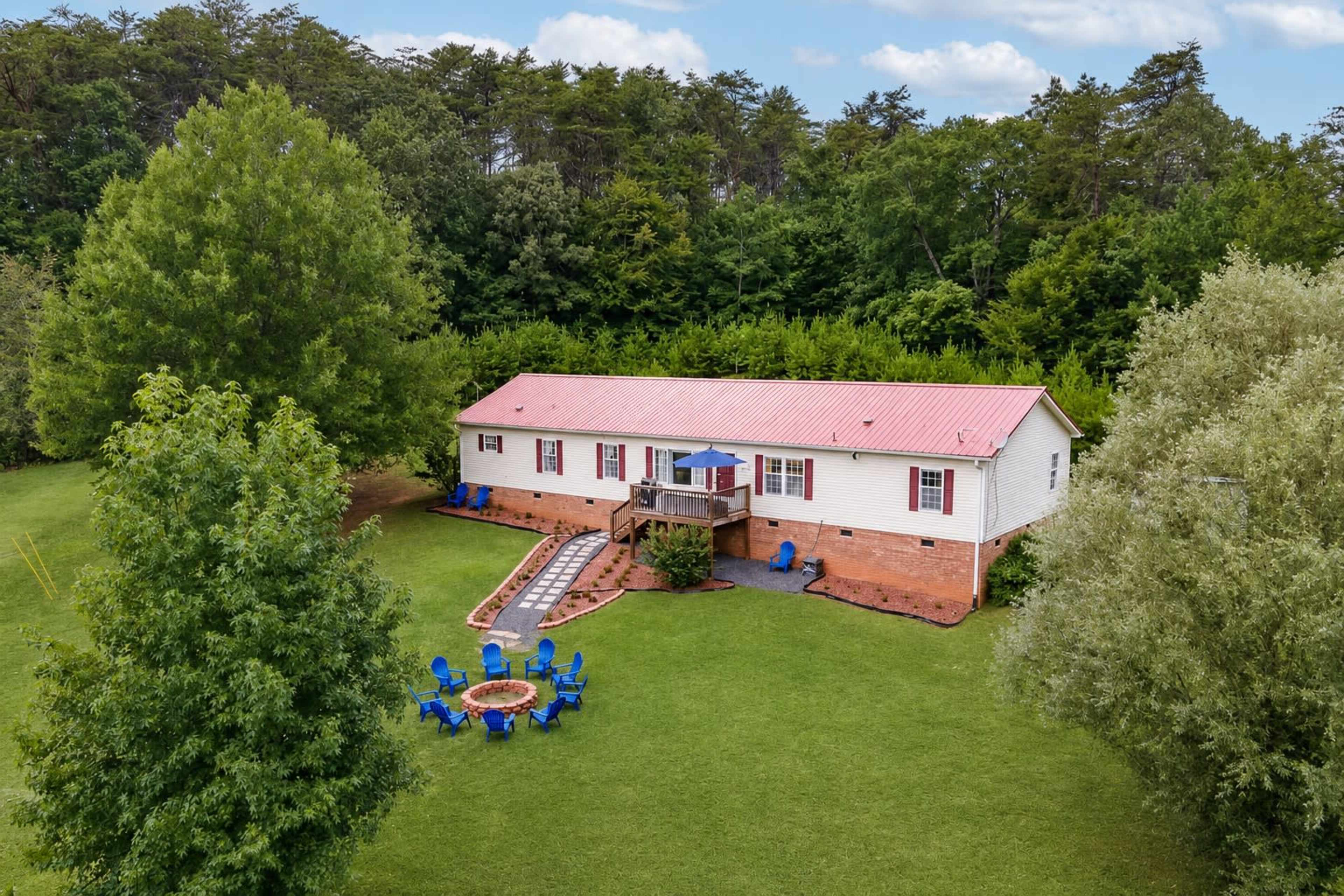 A single-story home with a red metal roof is situated on a grassy lot, surrounded by trees, with a patio and a circular arrangement of blue chairs around a fire pit.
