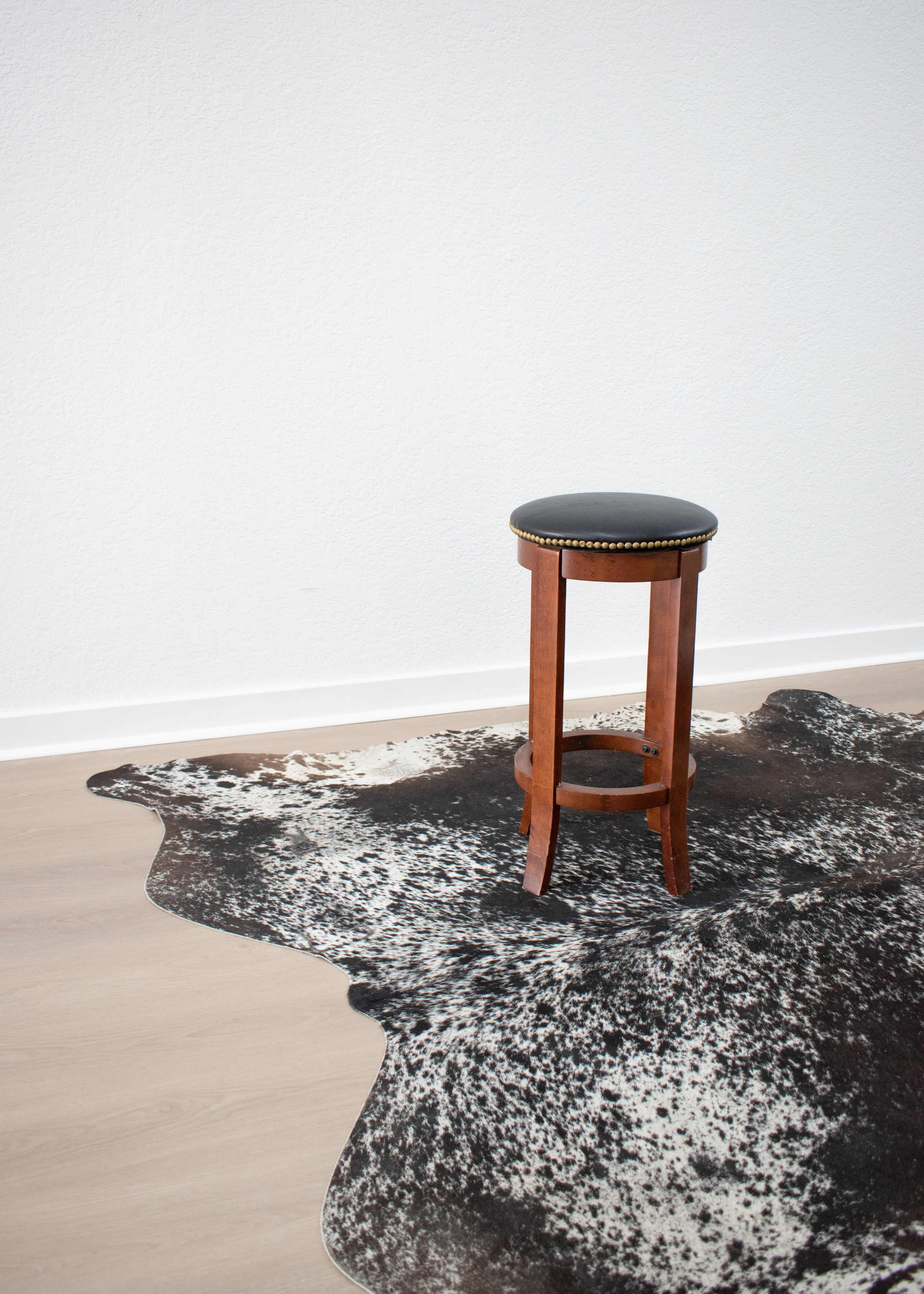 A wooden stool sits on a black and white patterned cowhide rug against a plain white wall.