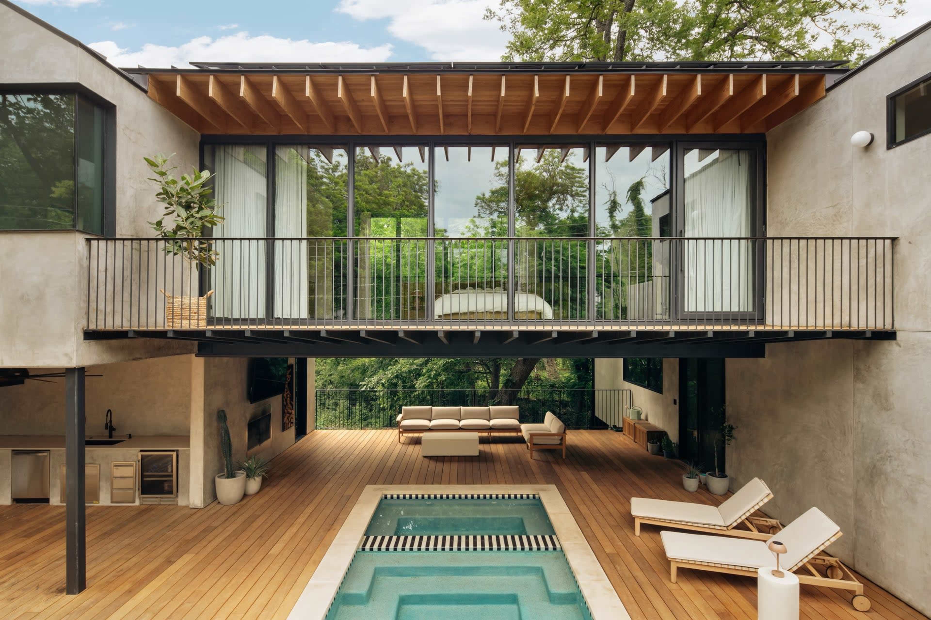 The image shows a modern house with a wooden deck, featuring a swimming pool, outdoor seating, and a large glass balcony overlooking a green landscape.