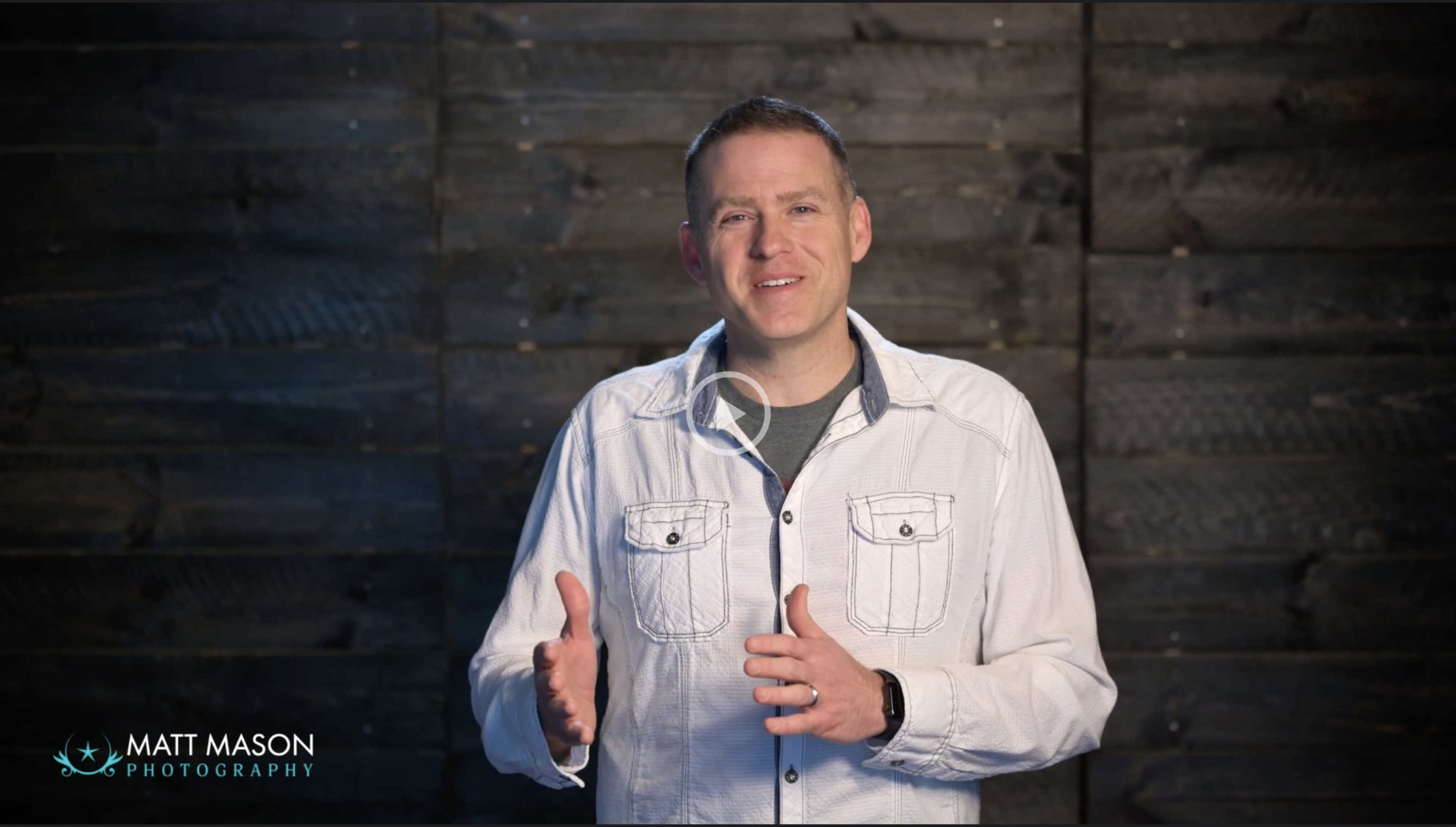A man stands in front of a wooden wall, gesturing with his hands as he speaks.