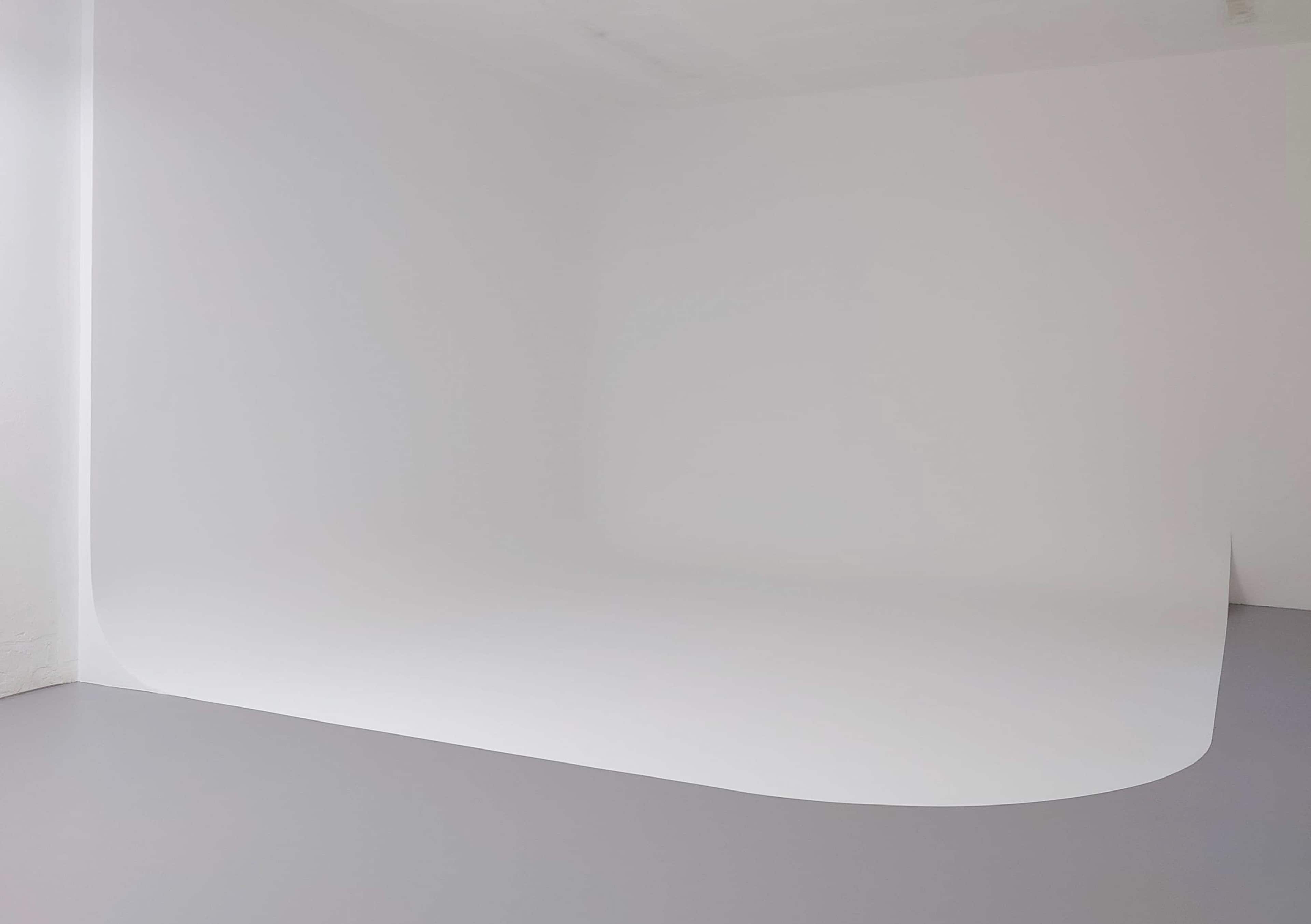 The image shows a minimalist studio space with a seamless white backdrop against a gray floor.