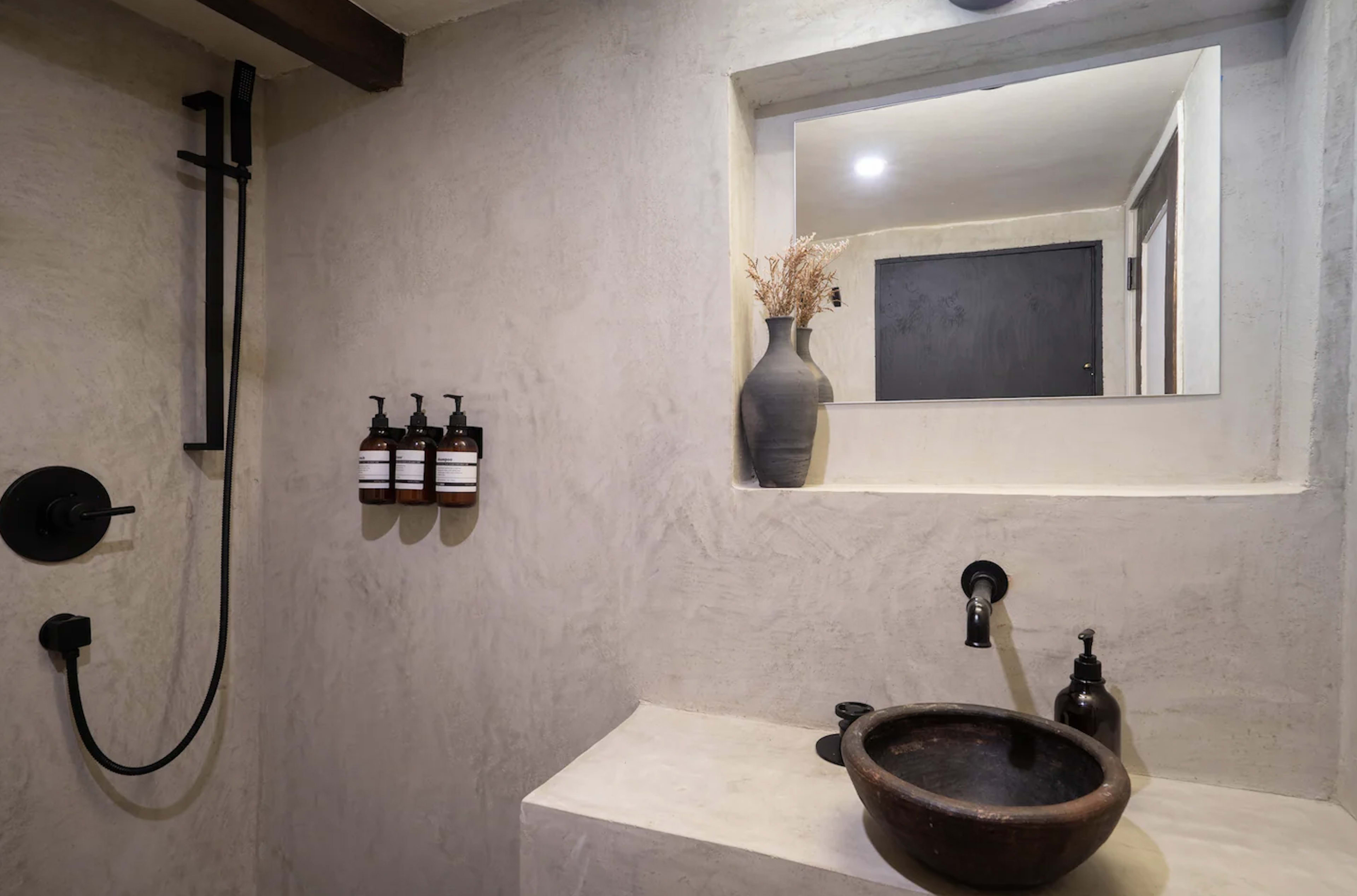 A minimalist bathroom featuring a stone sink, a black faucet, and a shower area with wall-mounted shampoo bottles and a decorative vase.