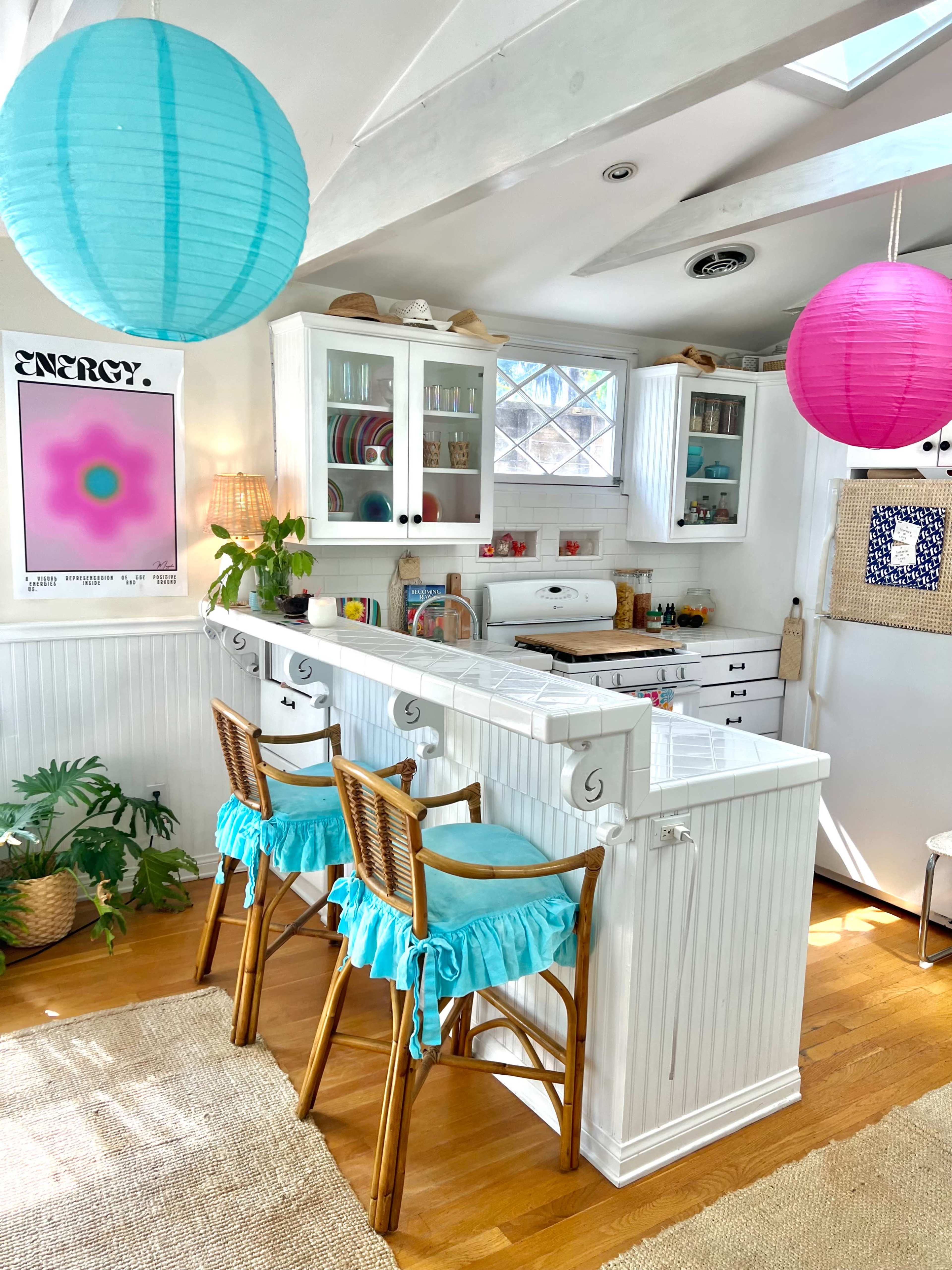 A bright kitchen features white cabinetry, bar stools with turquoise ruffles, and colorful paper lanterns hanging from the ceiling.