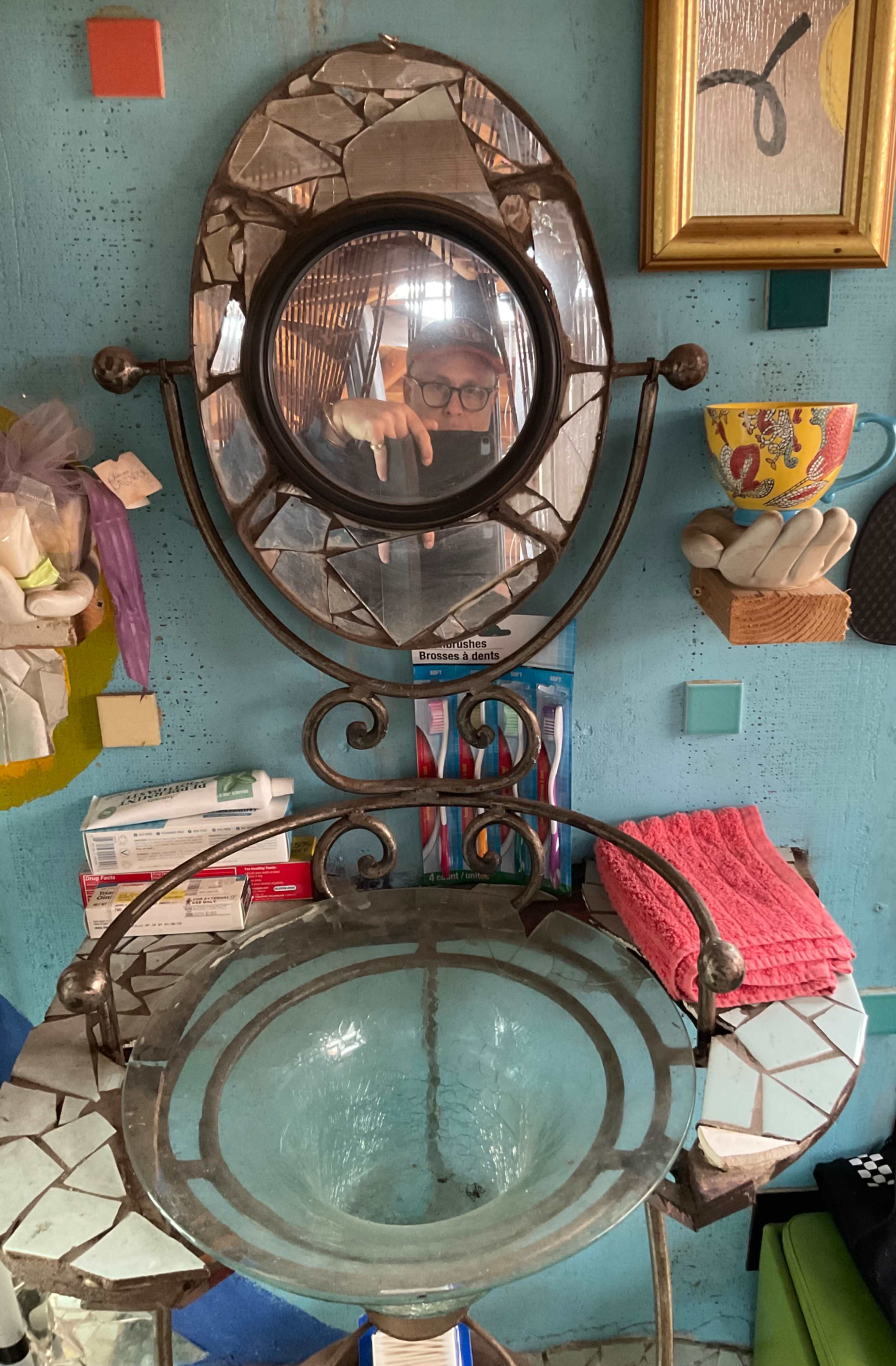 A decorative wall-mounted mirror with a circular shape is positioned above a glass basin, both set against a textured blue wall adorned with various items.