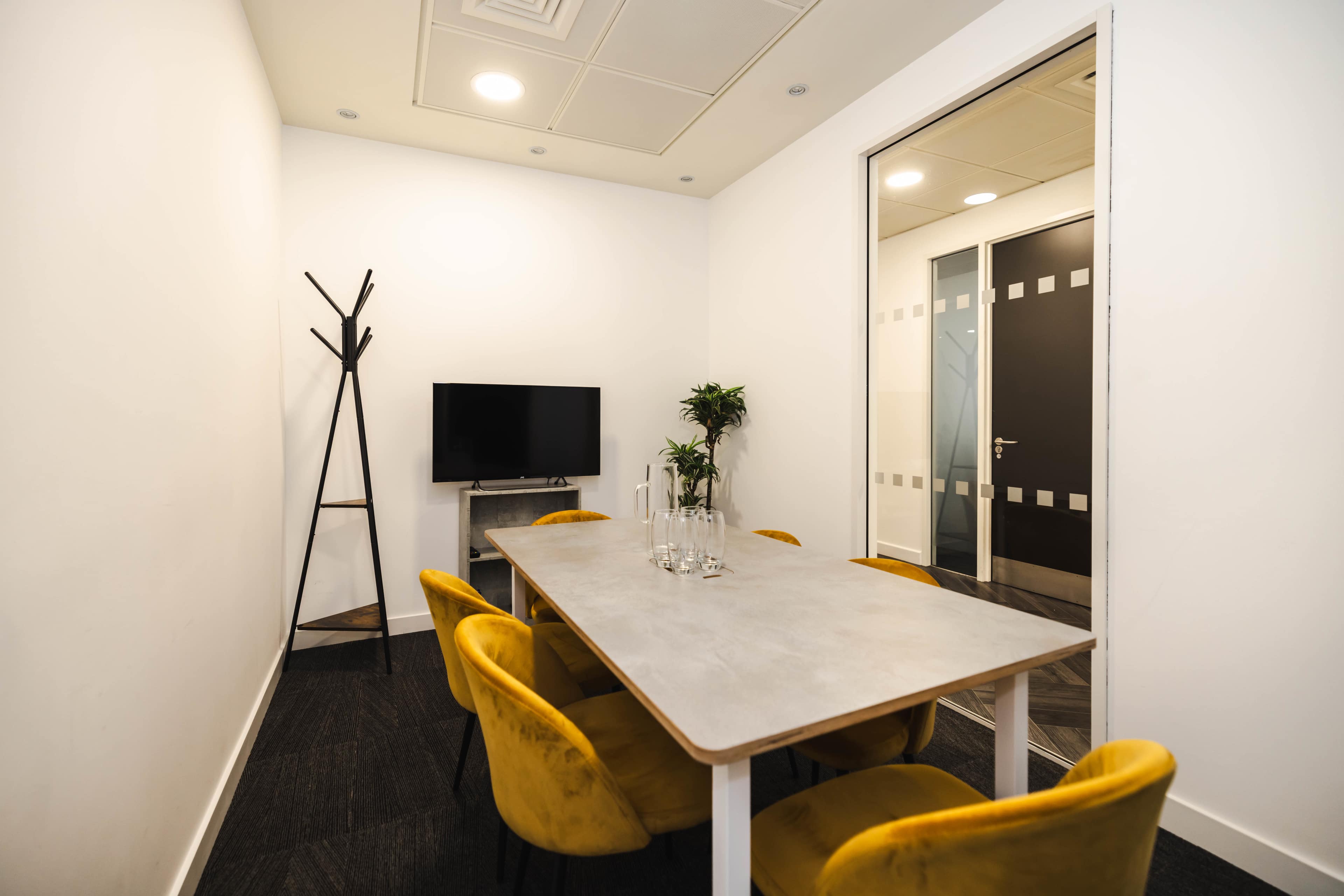A small meeting room features a gray table surrounded by yellow chairs, a television mounted on the wall, and a plant beside a coat rack.