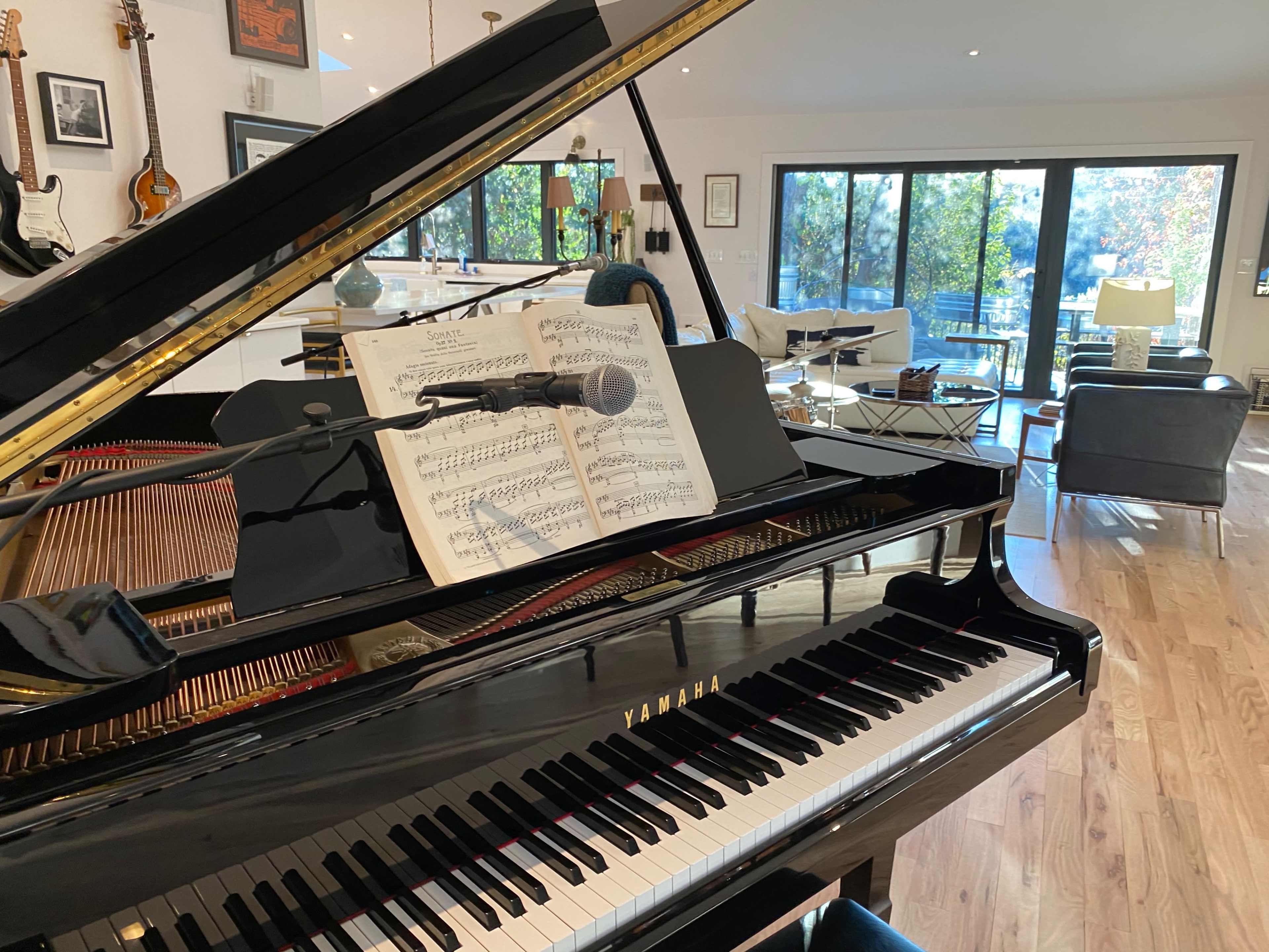 A grand piano with sheet music and a microphone is positioned in a bright room featuring large windows and a view of outdoor greenery.