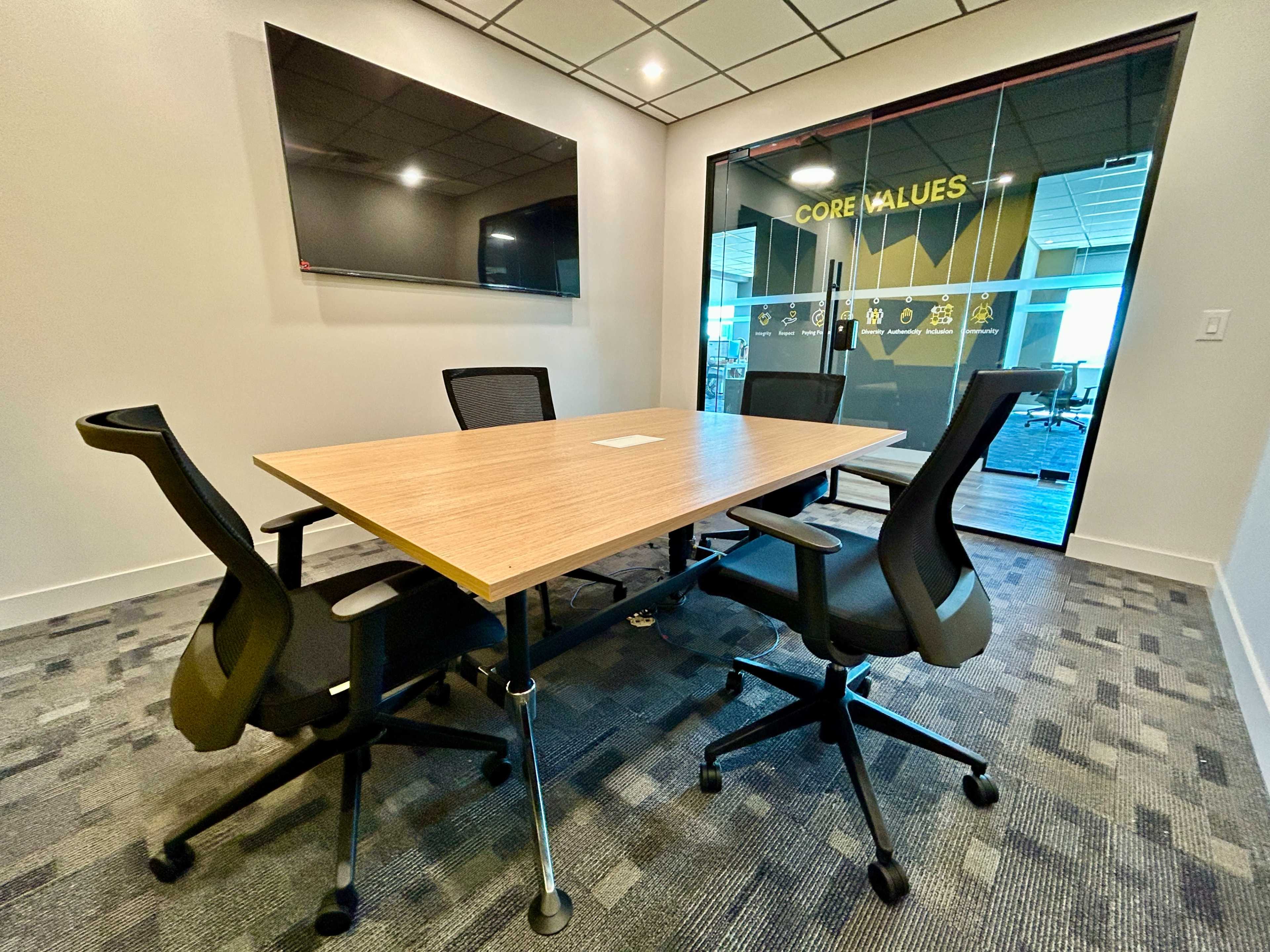 A small conference room features a wooden table surrounded by four black ergonomic chairs, with a screen mounted on one wall and glass doors displaying core values.
