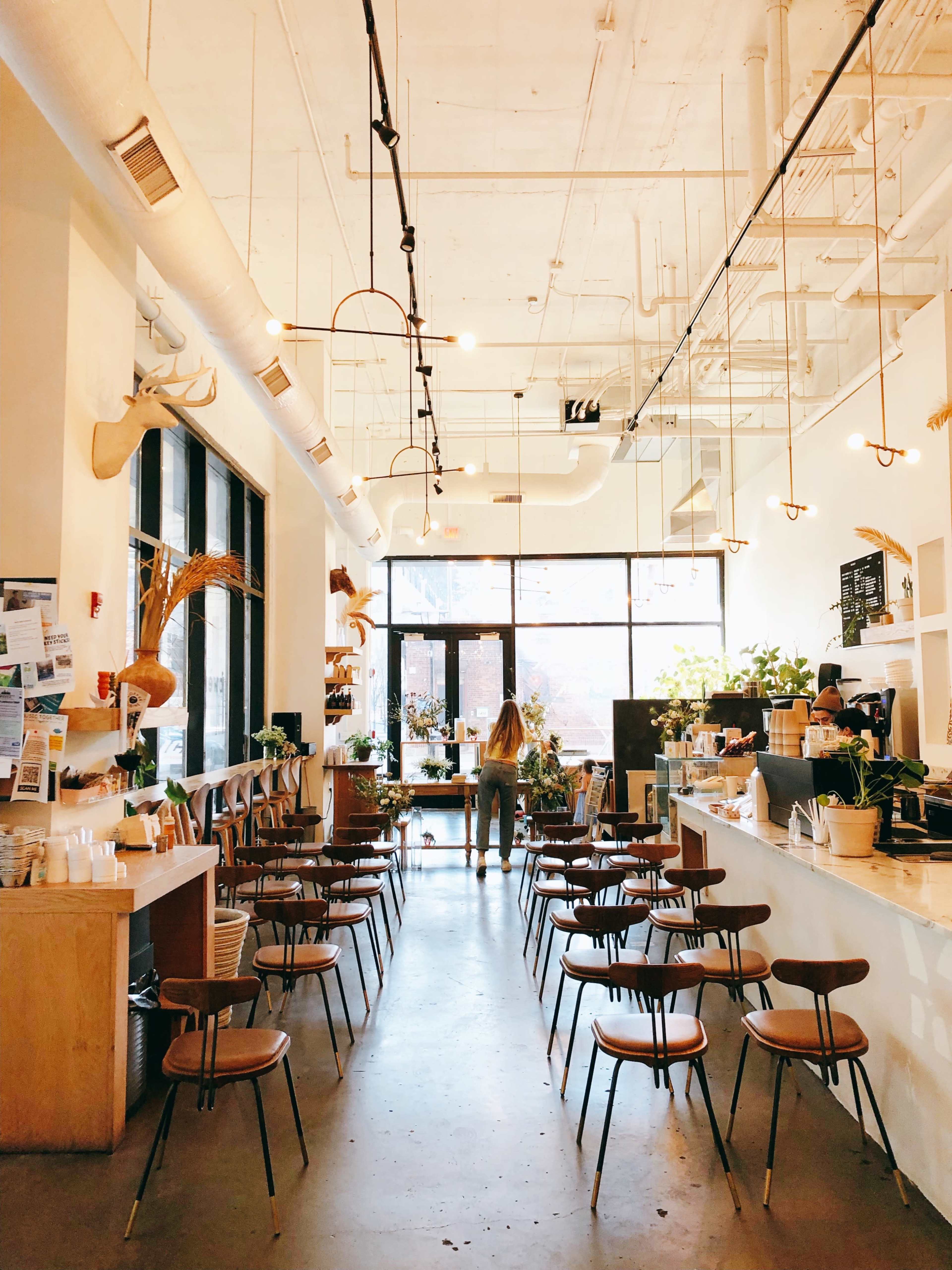 The image shows a spacious, modern café interior with wooden chairs arranged in rows and large windows letting in natural light.