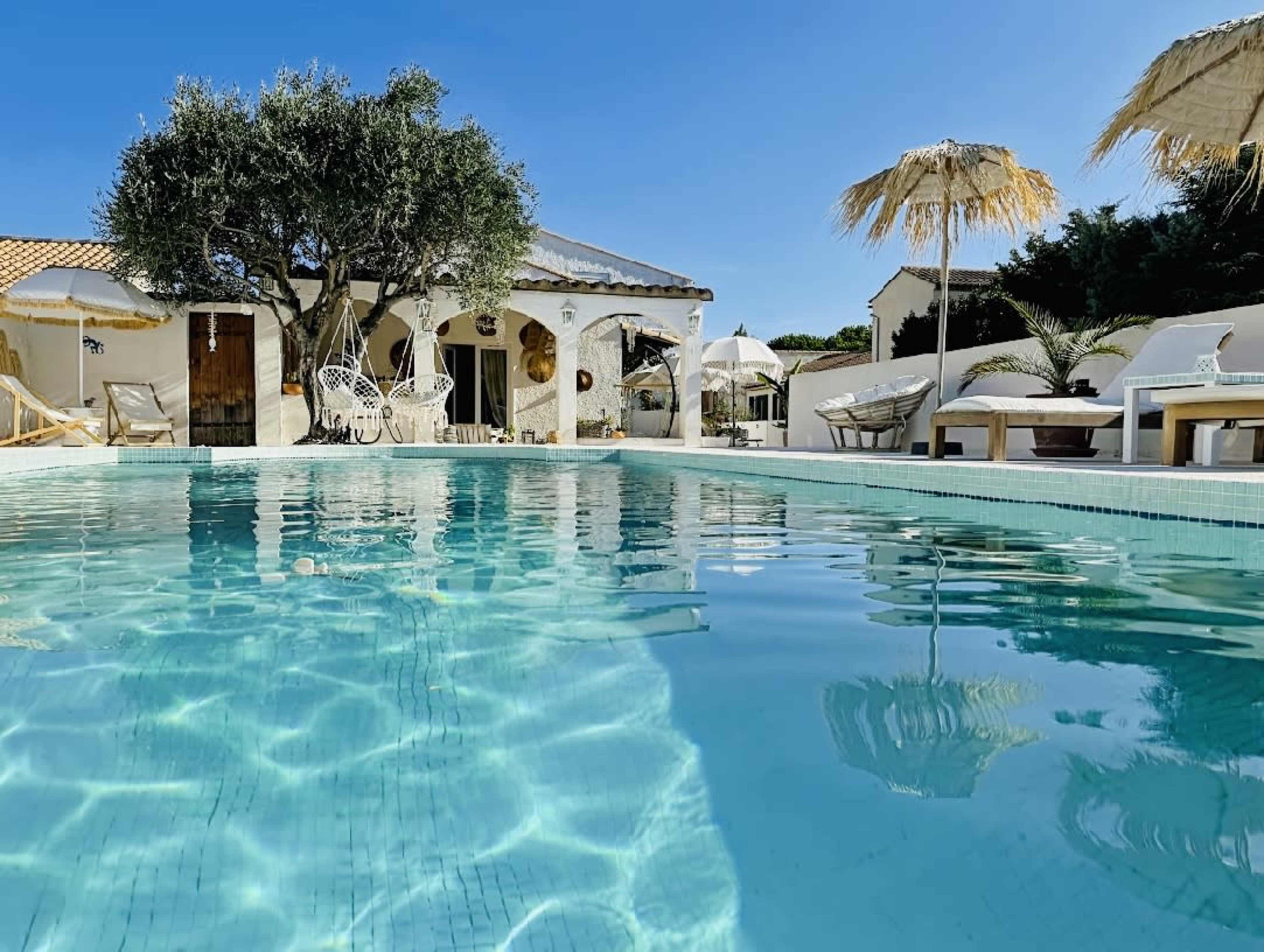 The image shows a clear swimming pool surrounded by a sunlit patio area with seating and umbrellas, adjacent to a white building with an olive tree in the background.