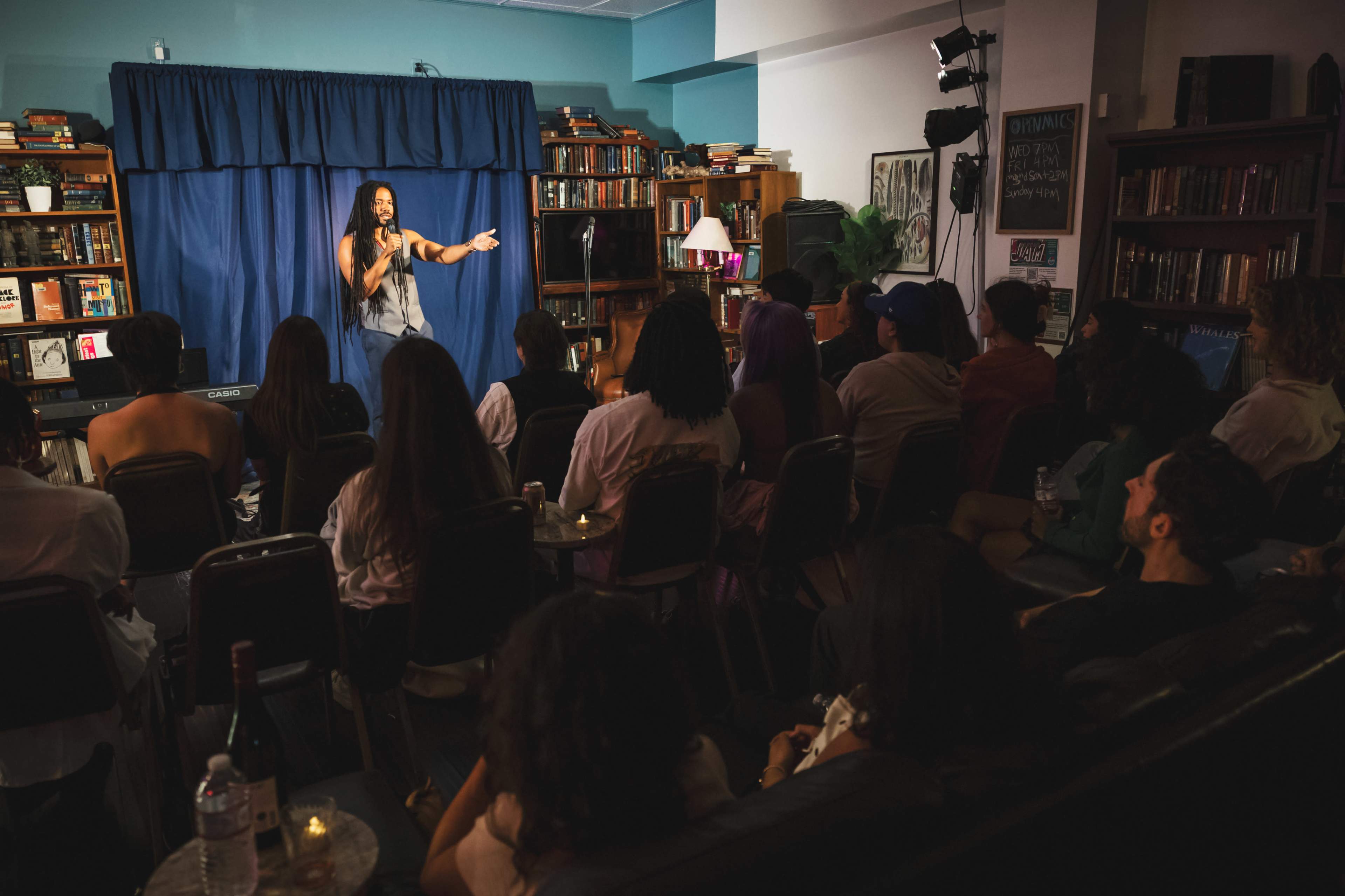 A performer stands on stage in front of an audience seated in a cozy, book-lined venue with blue curtains in the background.