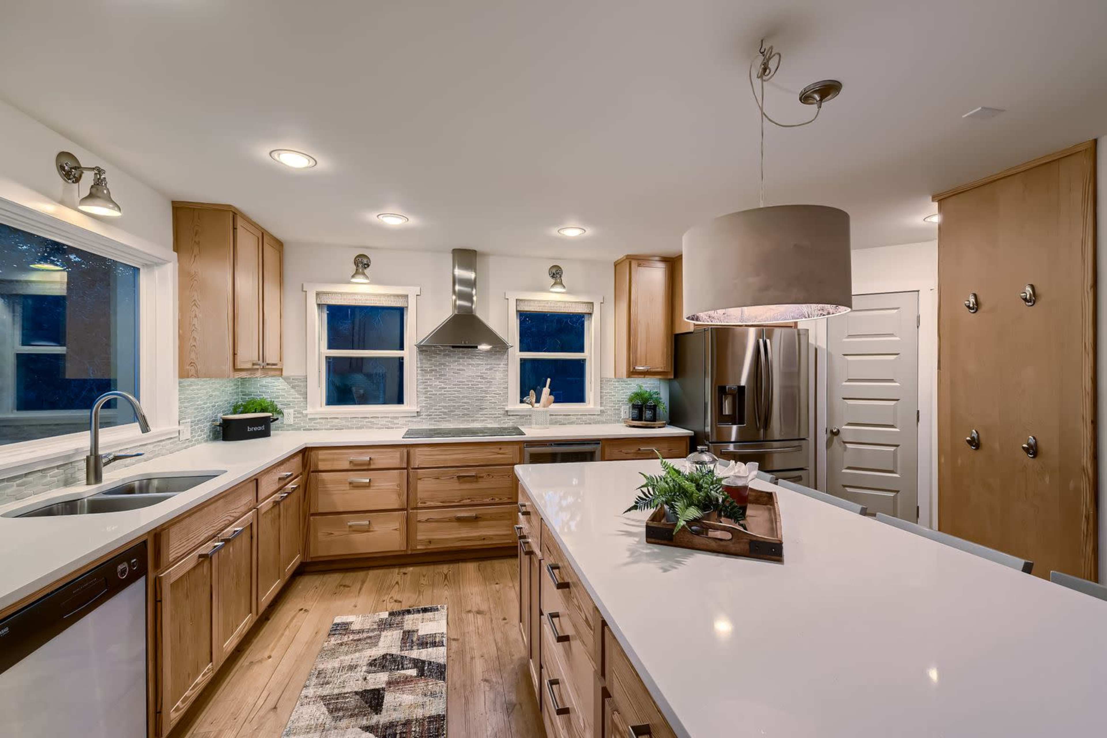 The image shows a modern kitchen featuring wooden cabinetry, stainless steel appliances, and a large island with a sink and decorative centerpiece.