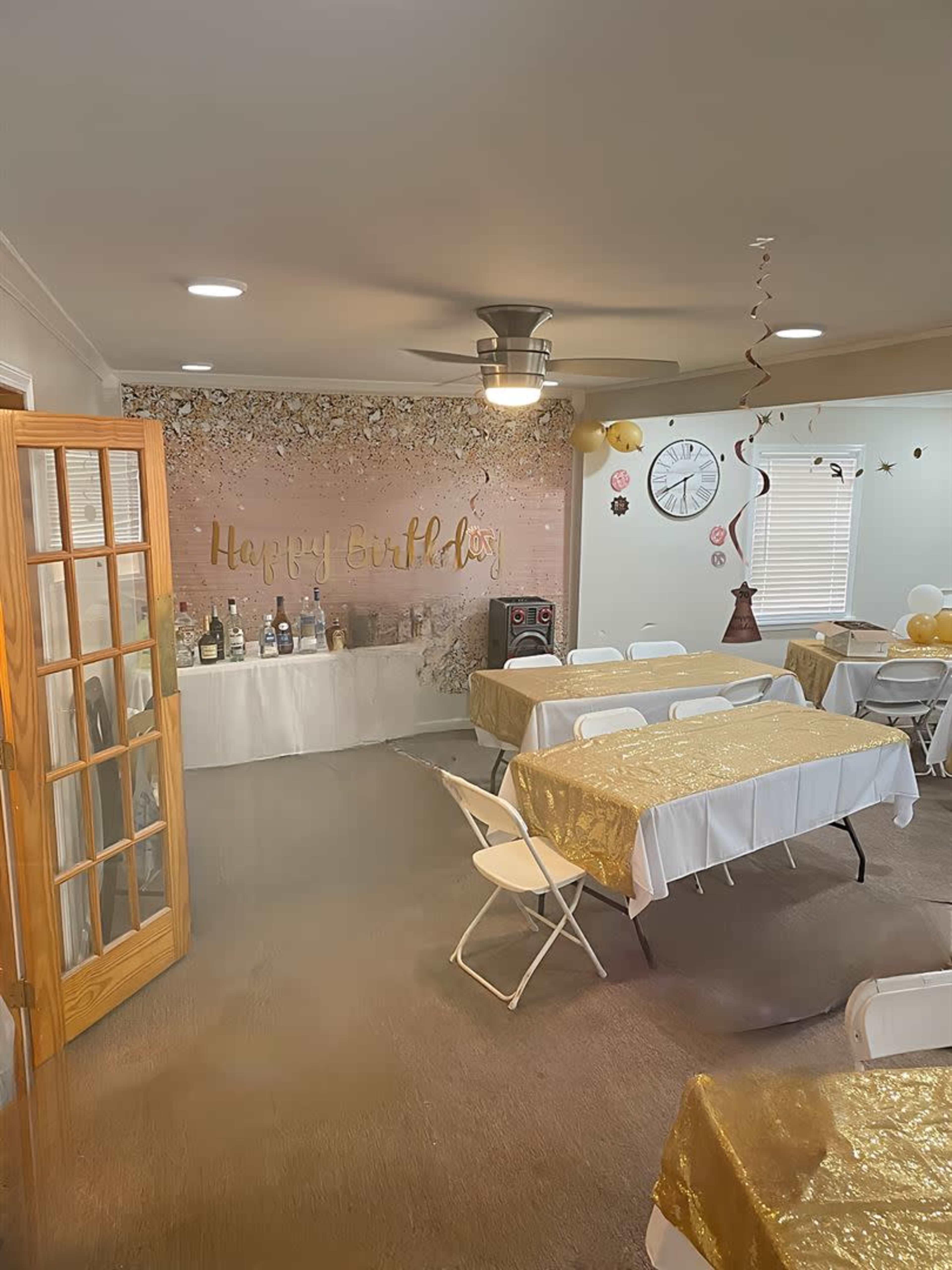 A decorated indoor space for a birthday celebration, featuring tables with gold tablecloths, a "Happy Birthday" sign on the wall, and various bottles arranged on a display table.