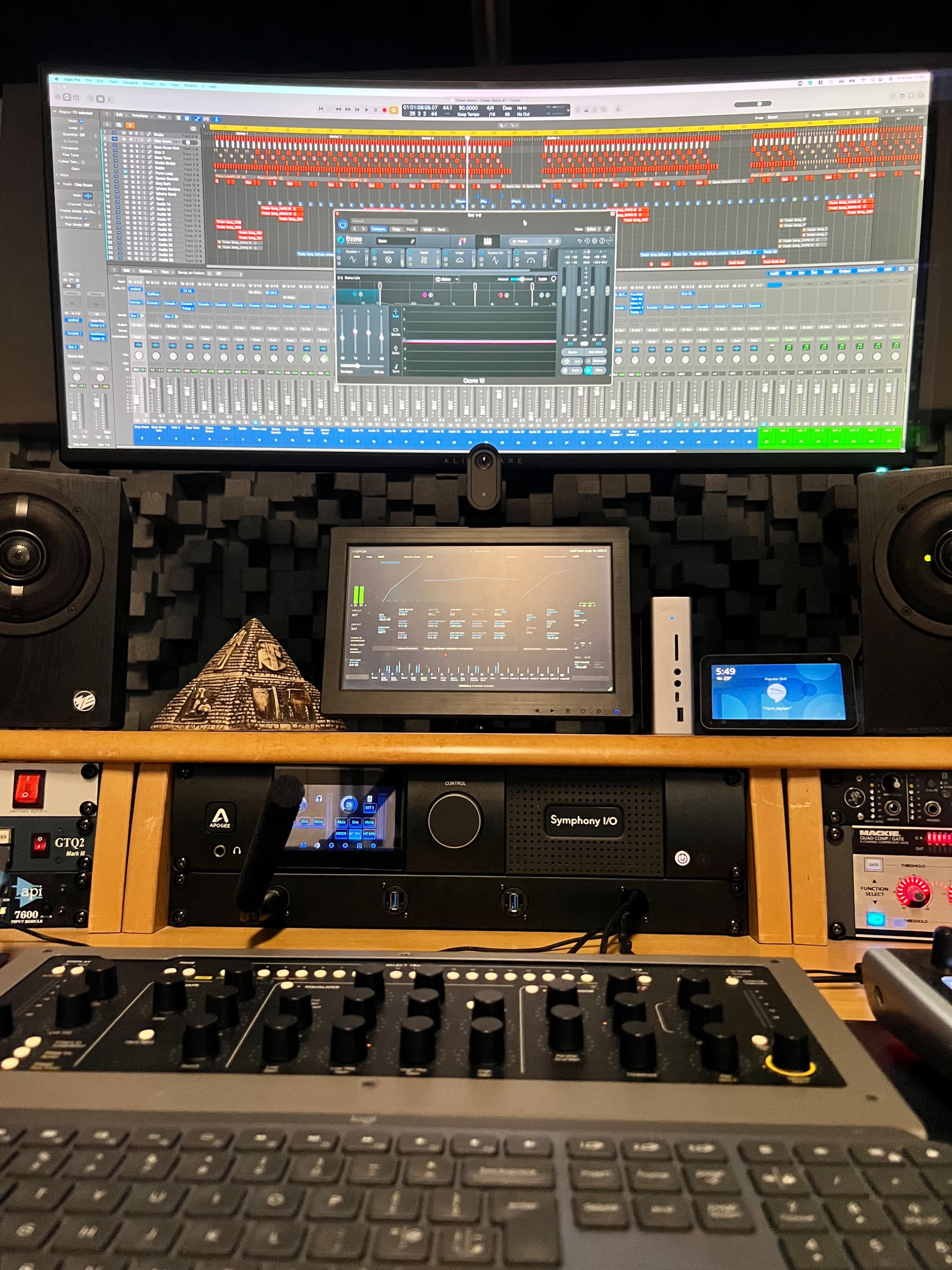 A music production studio with a large computer monitor displaying audio editing software and a control surface with various knobs and buttons.