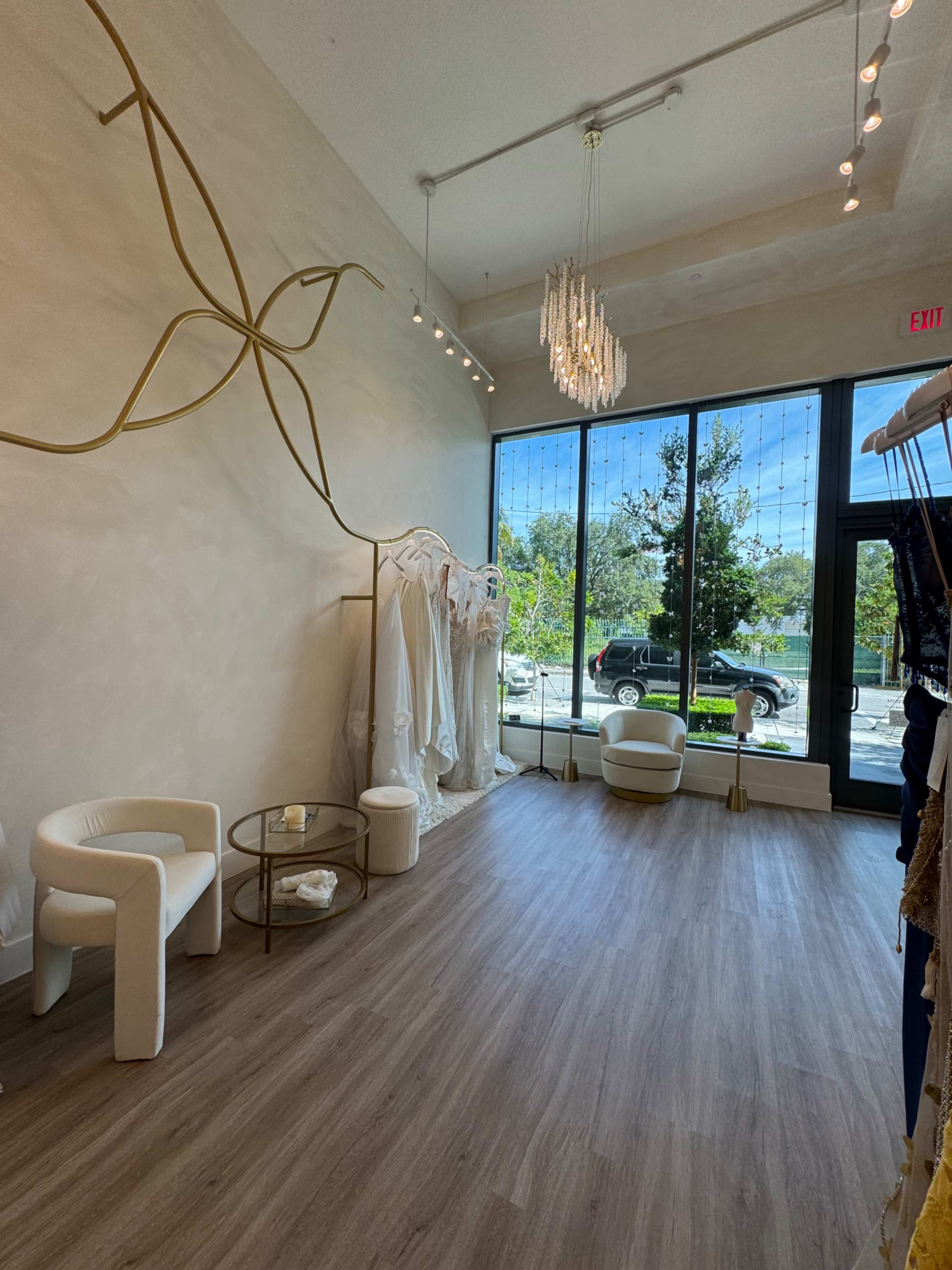 The interior of a boutique features a minimalist design with white clothing racks, two modern chairs, and a view of a sunny street through large glass windows.