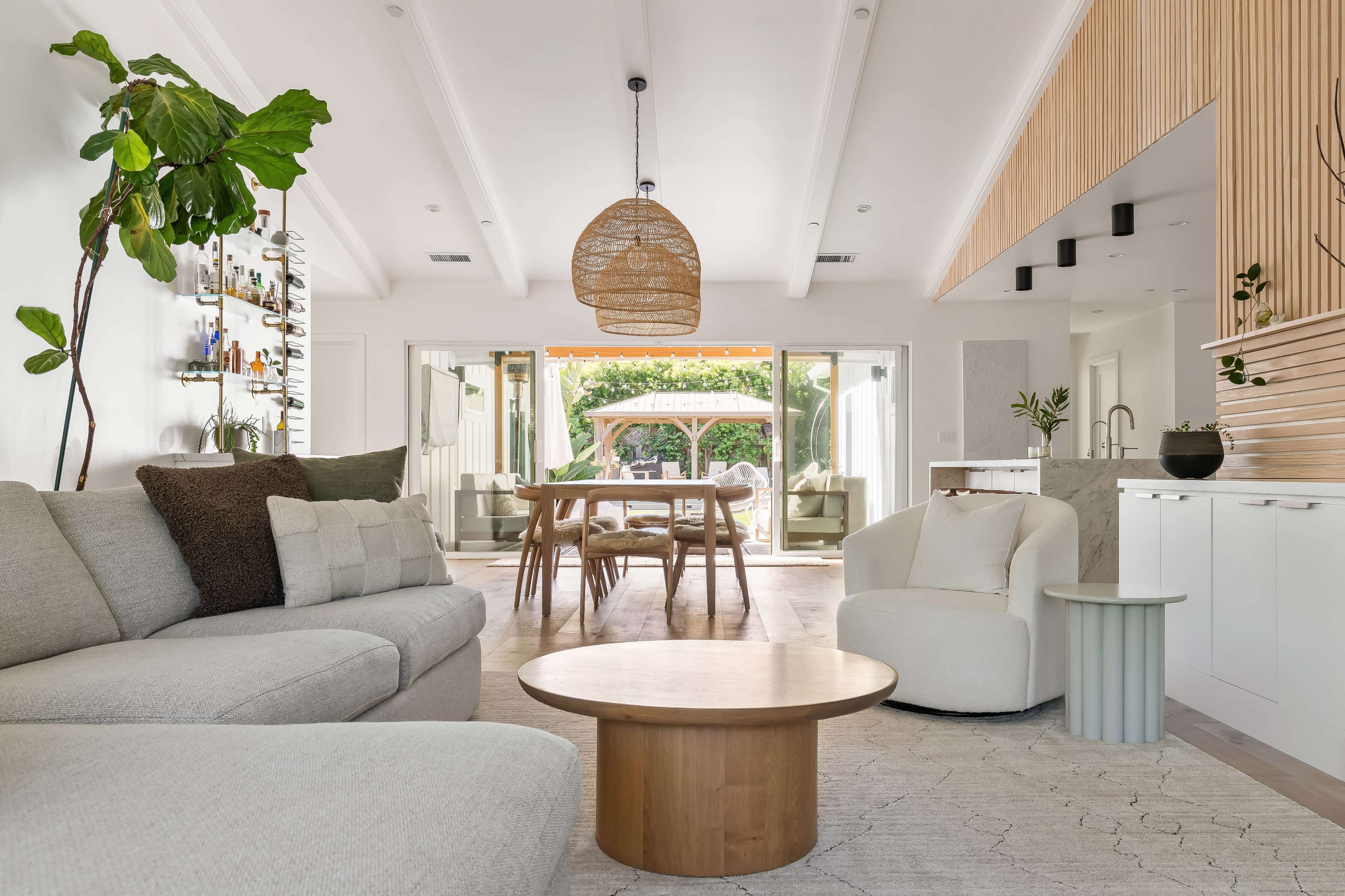 The image shows a modern living room with a light gray sectional sofa, a round wooden coffee table, and a dining area visible through glass doors that open to a garden.