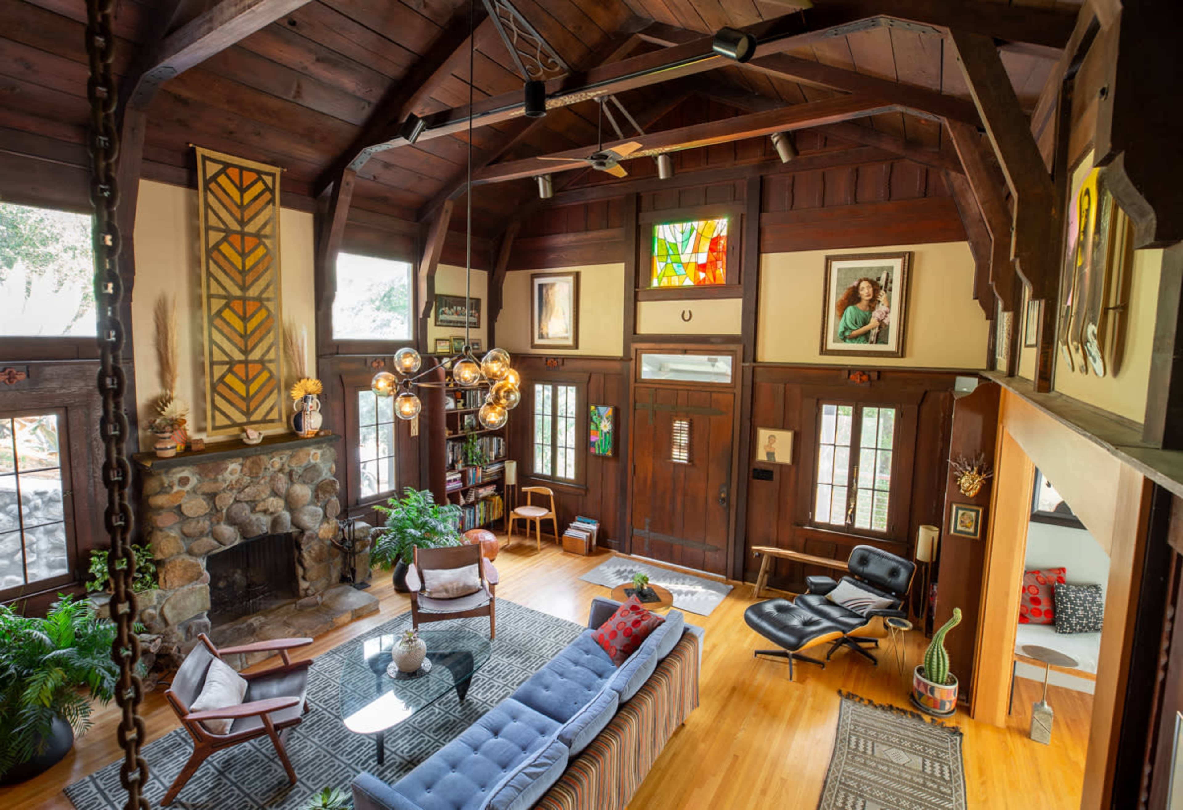 A spacious living room with a stone fireplace, wooden beams, and a mix of modern and vintage furniture, including a blue sofa and an art-decorated wall.