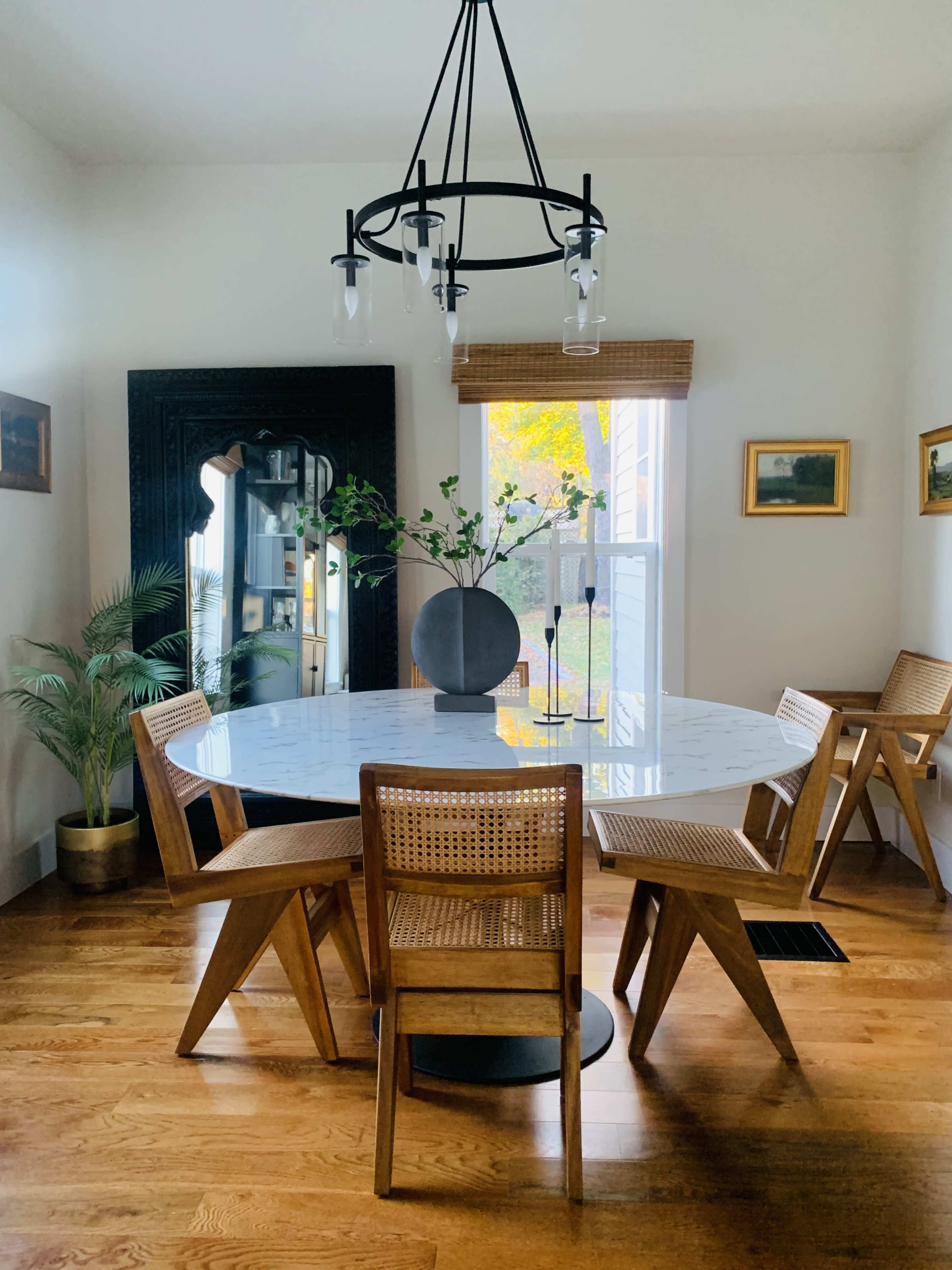 A round white dining table with wooden chairs is set in a bright room featuring a black mirror, framed pictures, and a plant.