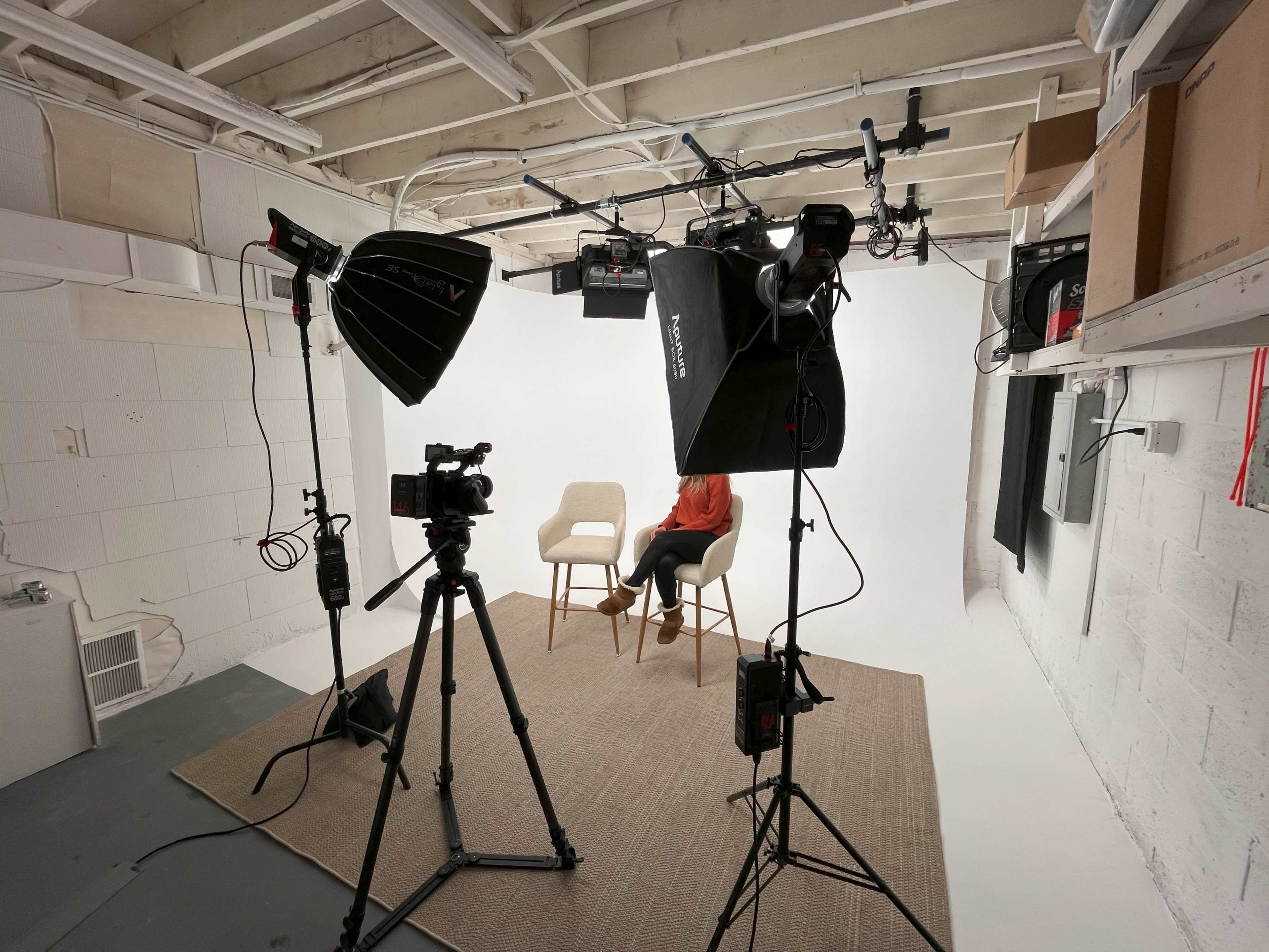 A studio setup features two chairs on a rug, with multiple lights and cameras positioned to capture a seated individual against a white backdrop.