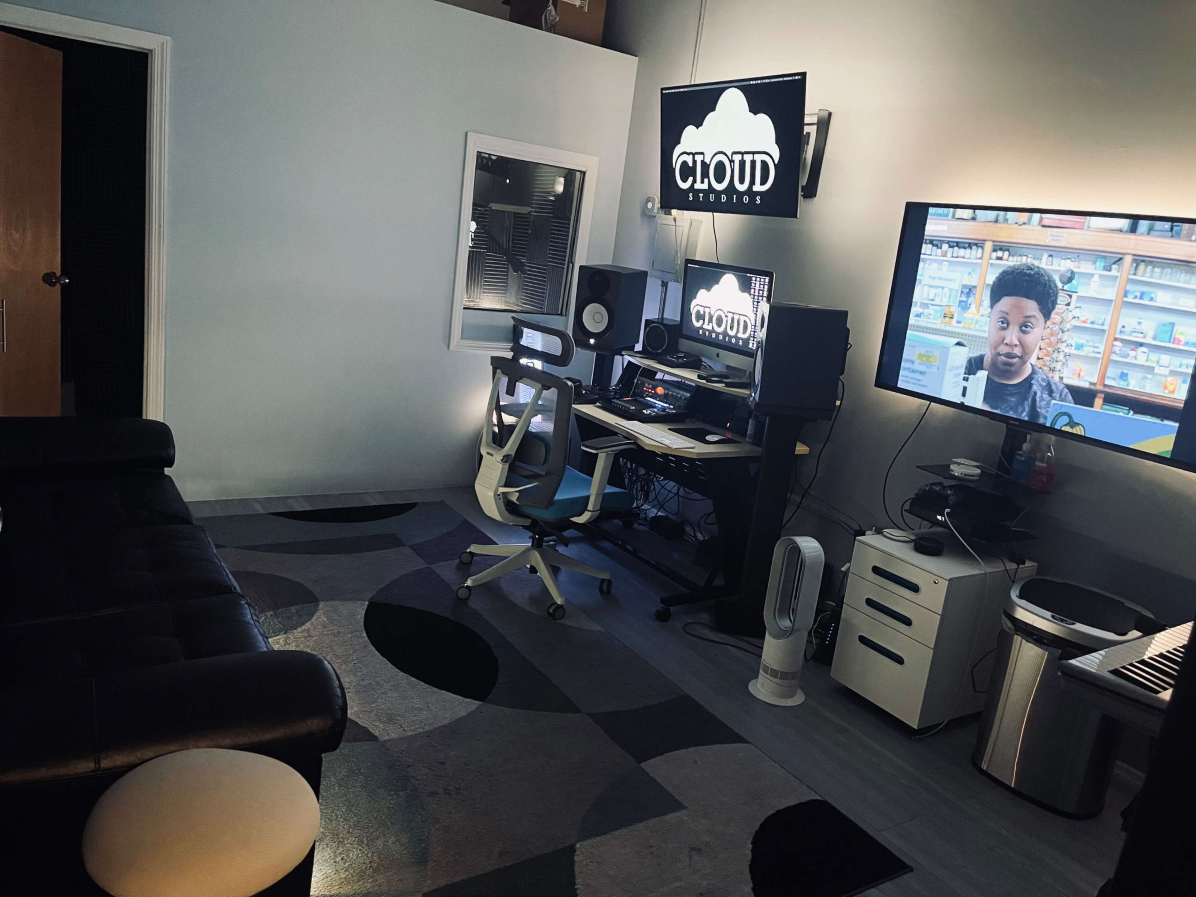 A recording studio with a computer desk, a large screen displaying a video, and a black couch in a room with modern decor.