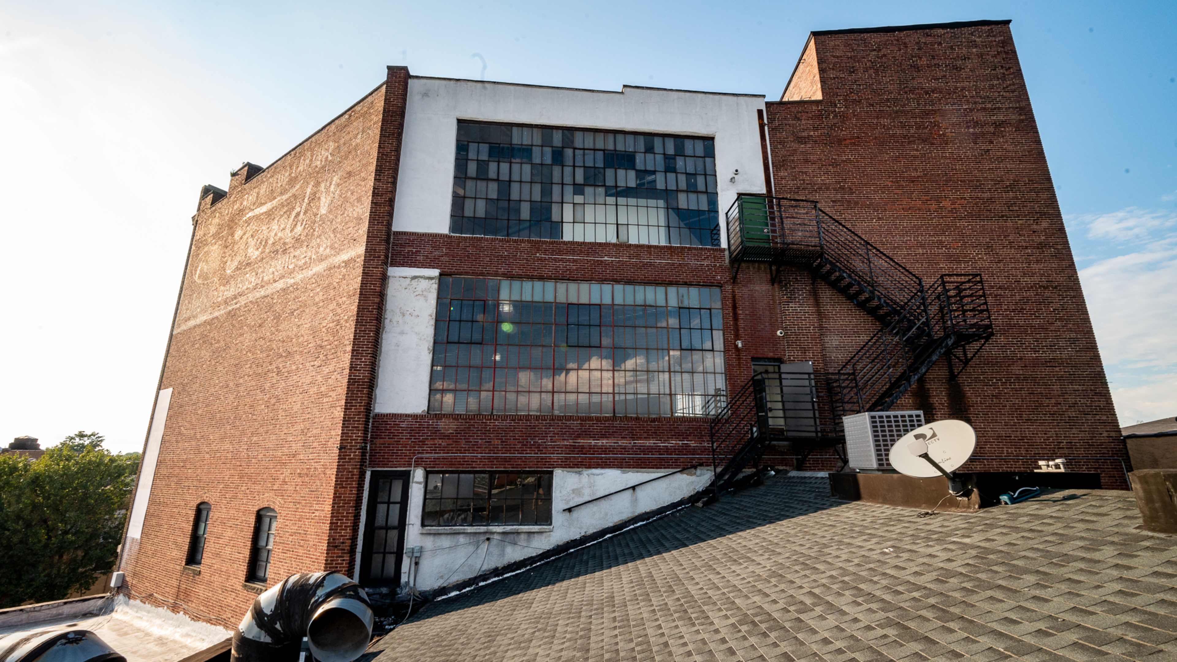 The rear view of a brick building with large windows and an external fire escape, situated adjacent to a sloped roof.