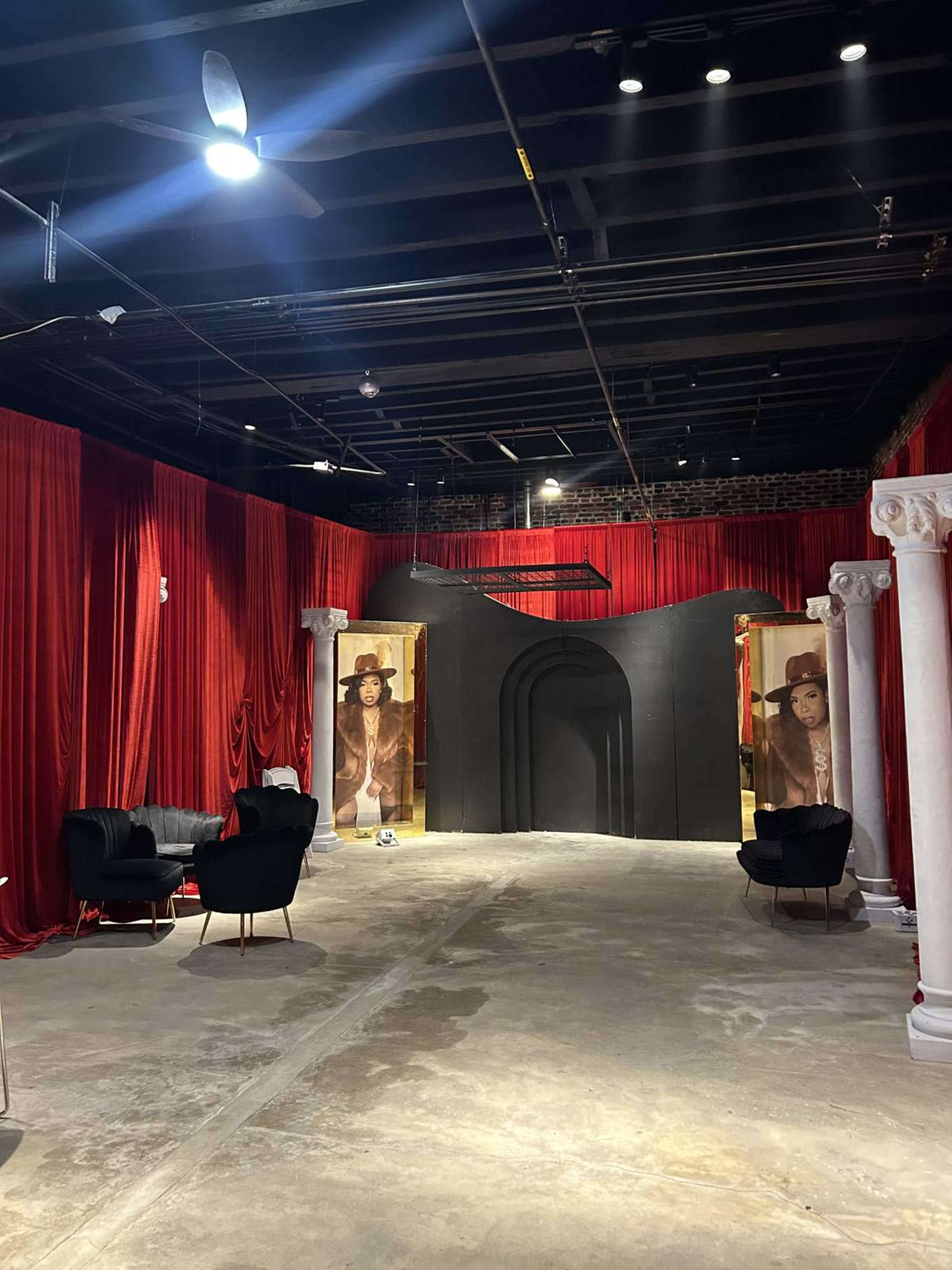 The image shows an empty room with red drapery, two large black archways framed by white columns, and a couple of black chairs arranged in the space.