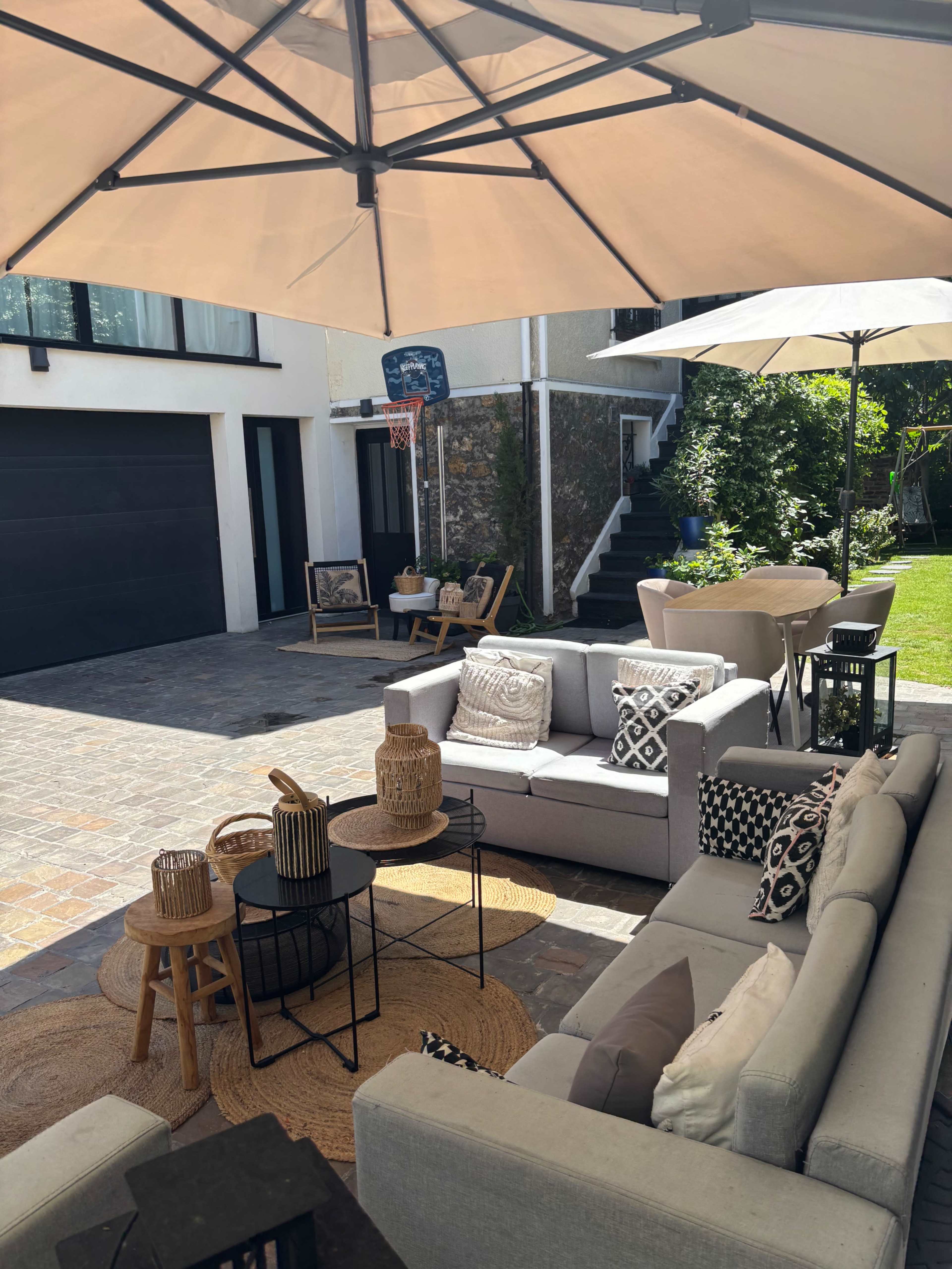 The image shows a spacious outdoor seating area featuring gray sofas, a round table, and various decorative elements, all under large umbrellas in a well-maintained backyard.