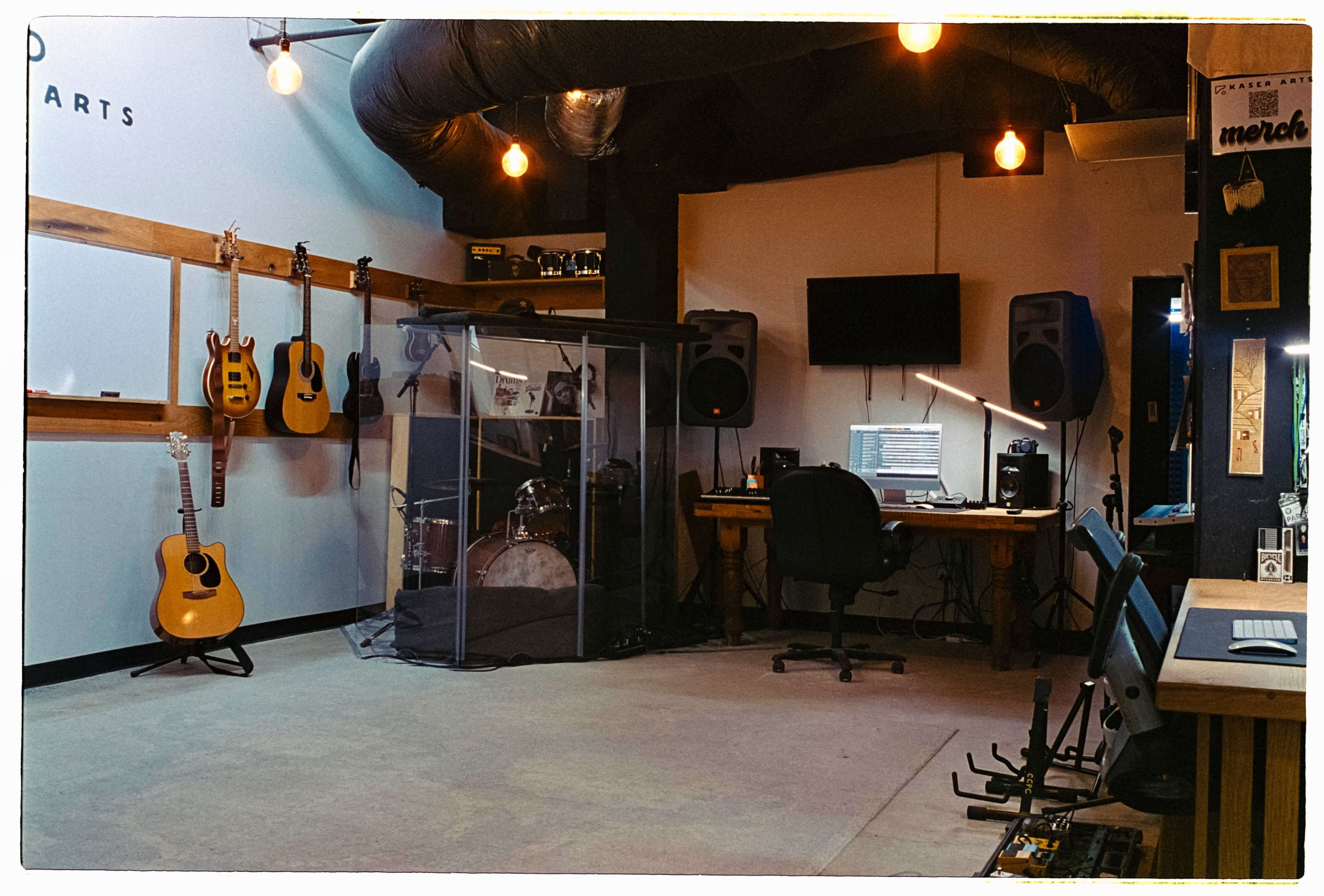 The image shows a music studio featuring guitars on the wall, a drum set enclosed in glass, and a workspace with a computer and audio equipment.