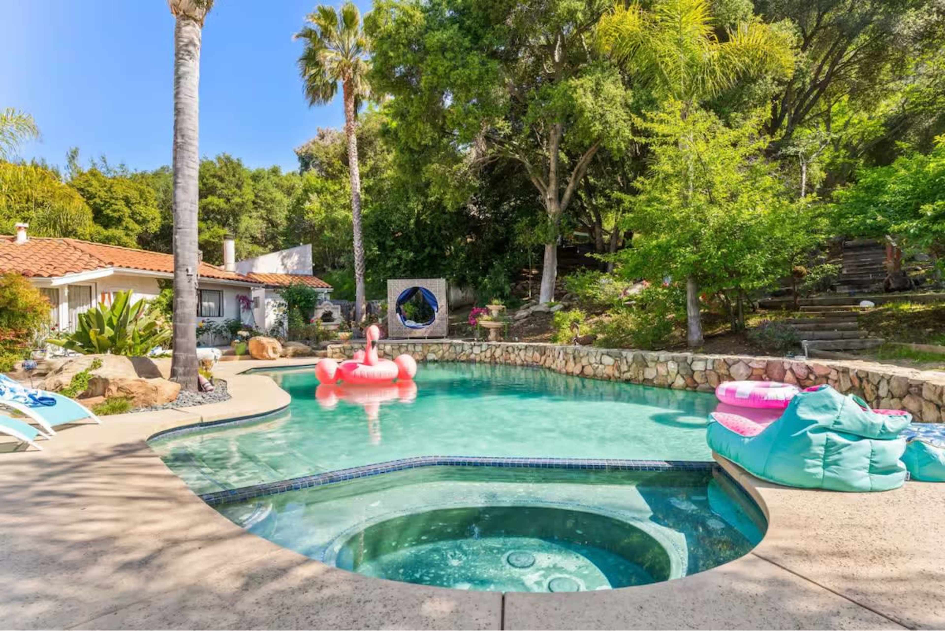 The image shows a backyard pool area with a hot tub, colorful inflatable float, and lush vegetation surrounding the space.