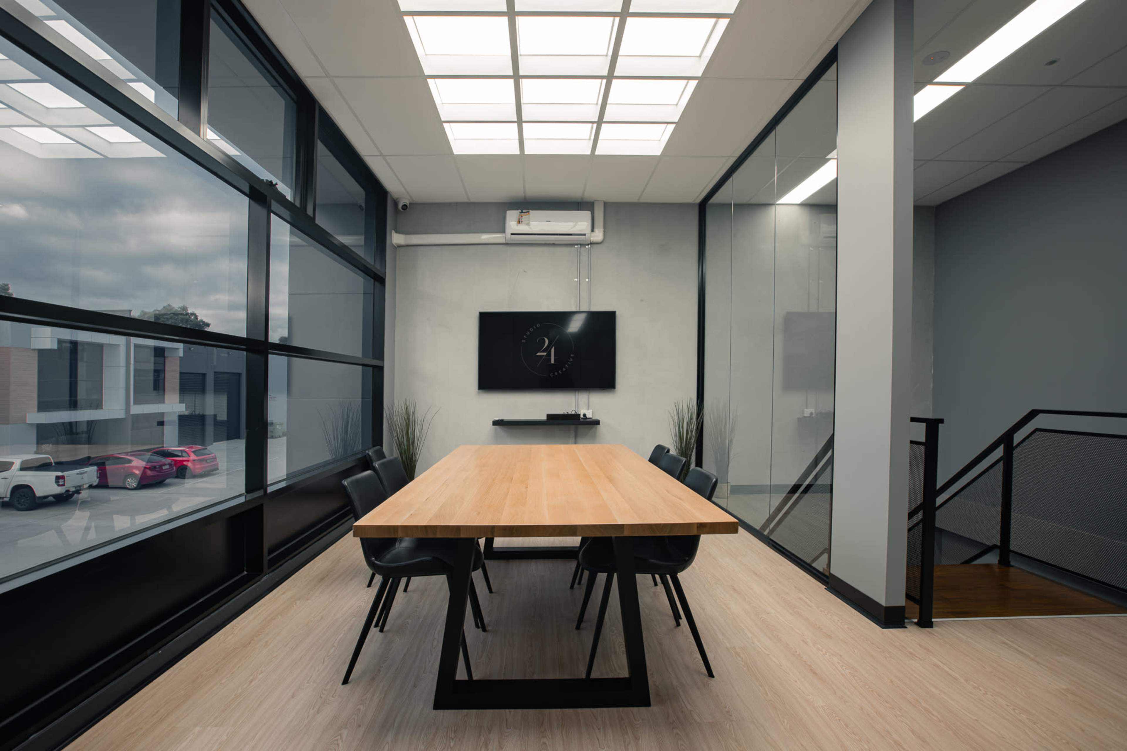 A modern conference room features a large wooden table surrounded by black chairs, with glass walls and a TV mounted on the wall, illuminated by natural light from the windows.