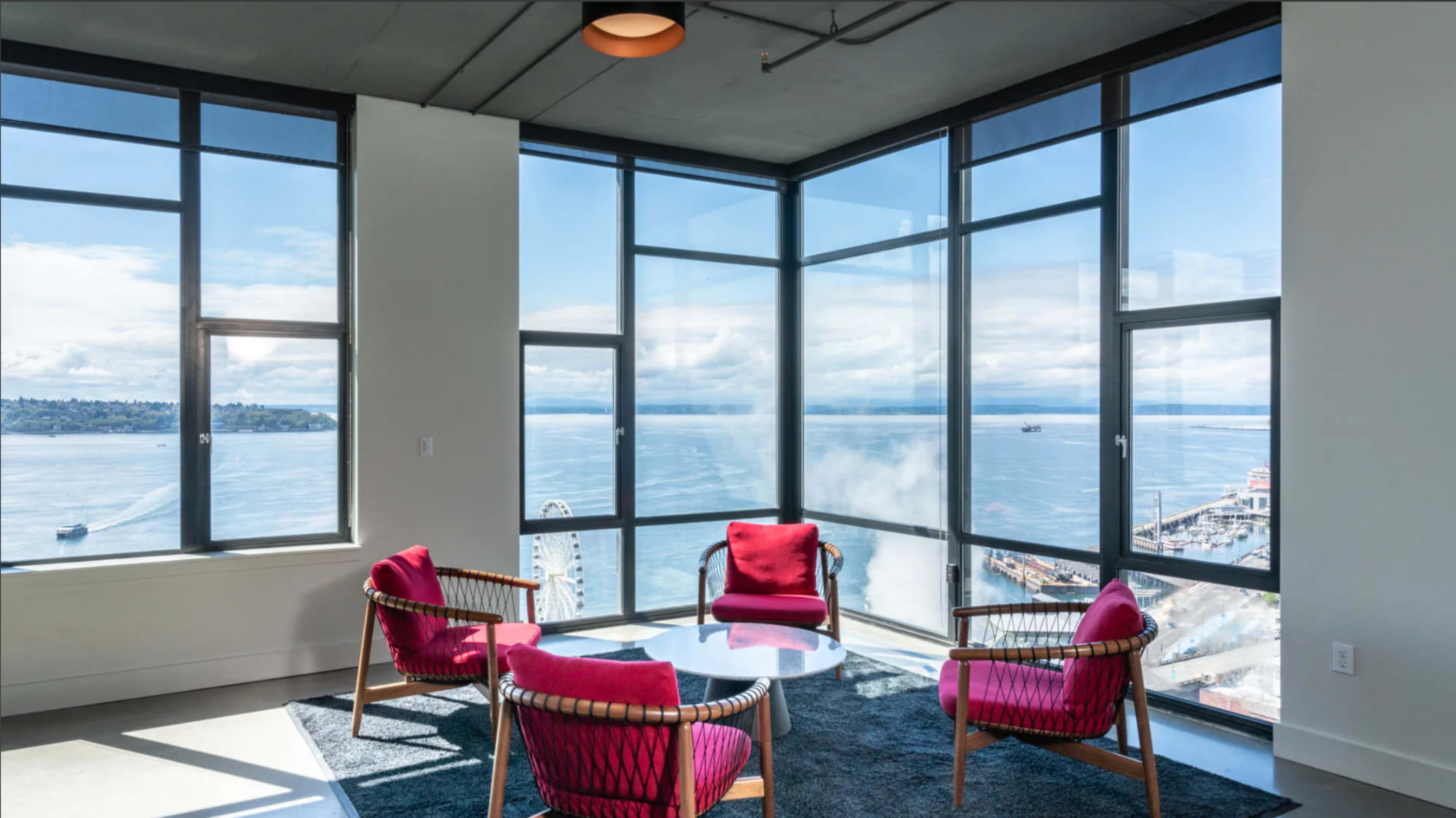 The image shows a modern sitting area with large windows offering a view of a body of water and a cityscape, featuring four chairs and a round table.