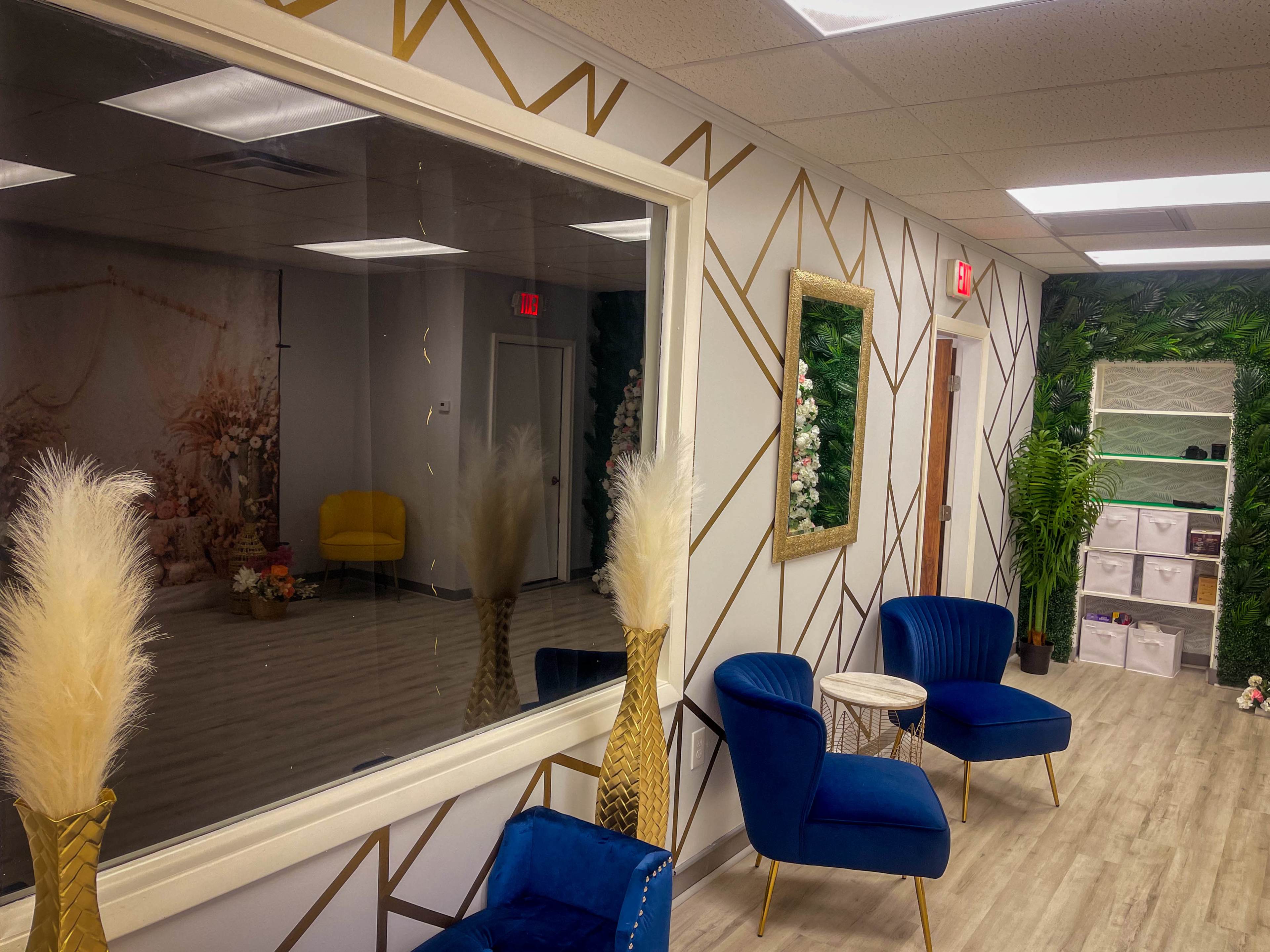 A modern waiting area featuring two blue chairs, a small table, decorative plants, and a large window overlooking a stylized backdrop.