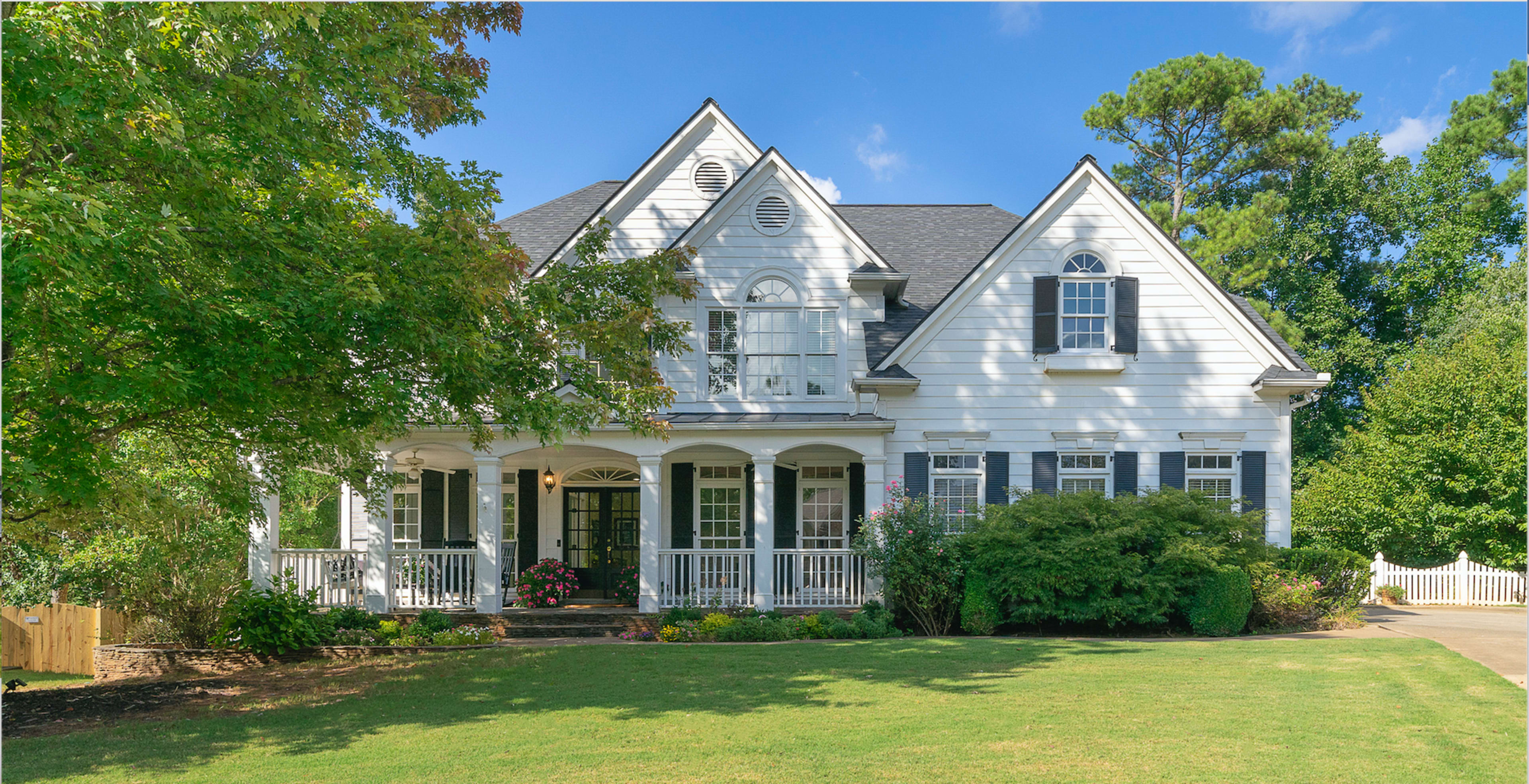 A two-story white house with black shutters and a front porch is surrounded by lush greenery and trees.