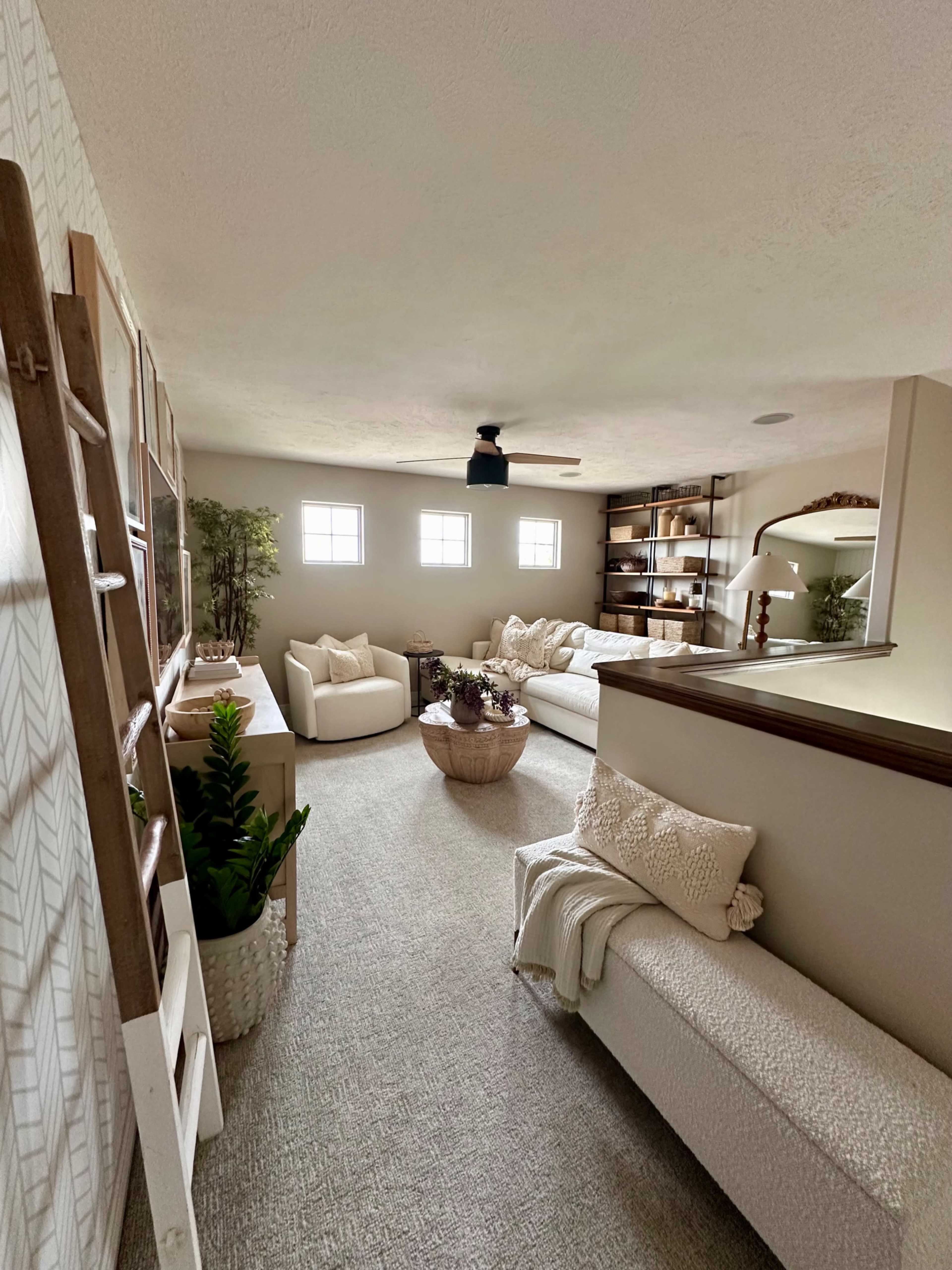 The image shows a cozy living area with two sofas, a small round table, and shelves, illuminated by natural light coming through nearby windows.