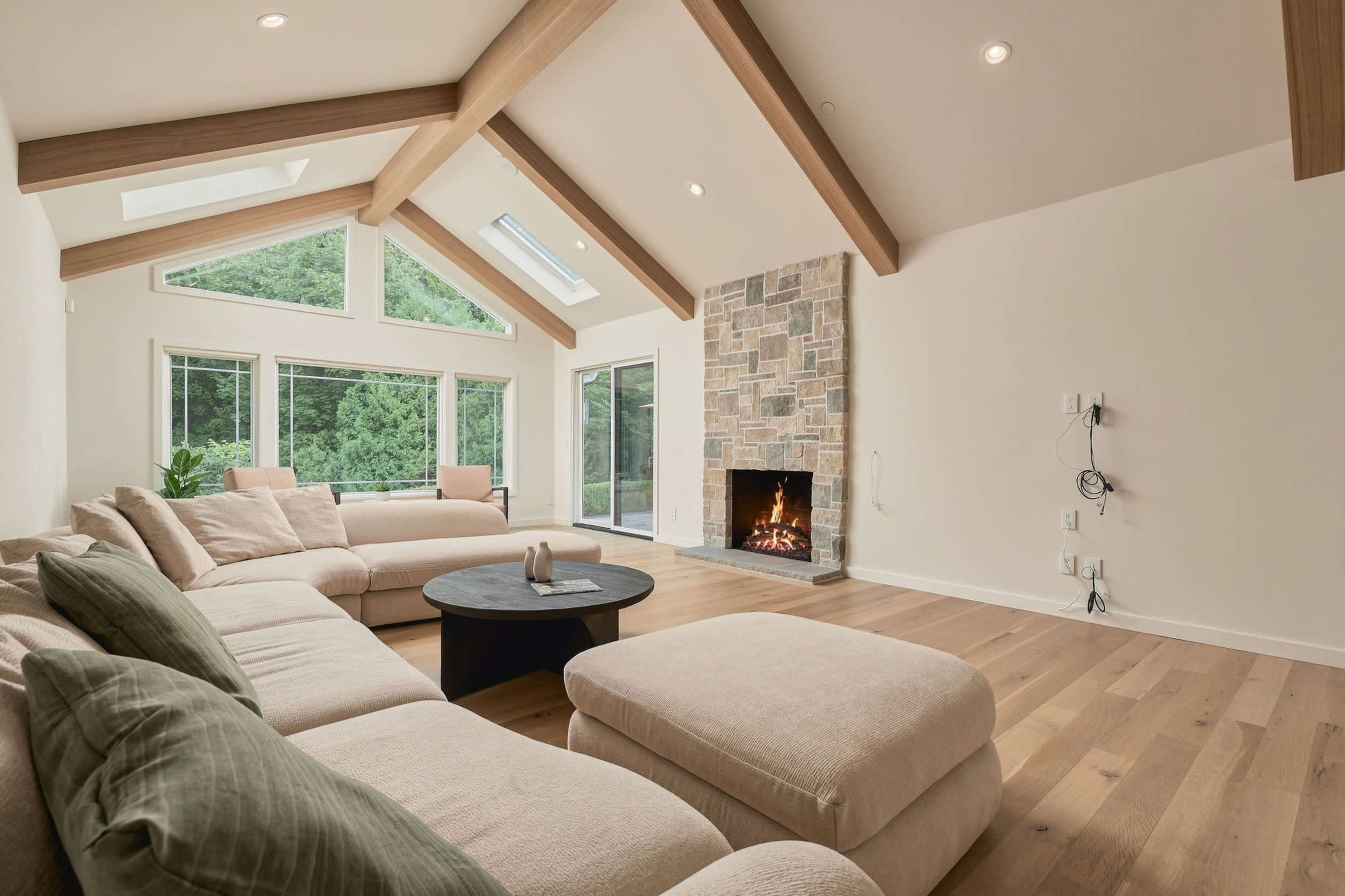 The image shows a spacious living room with a stone fireplace, large windows, and a comfortable sectional sofa arranged around a round coffee table.