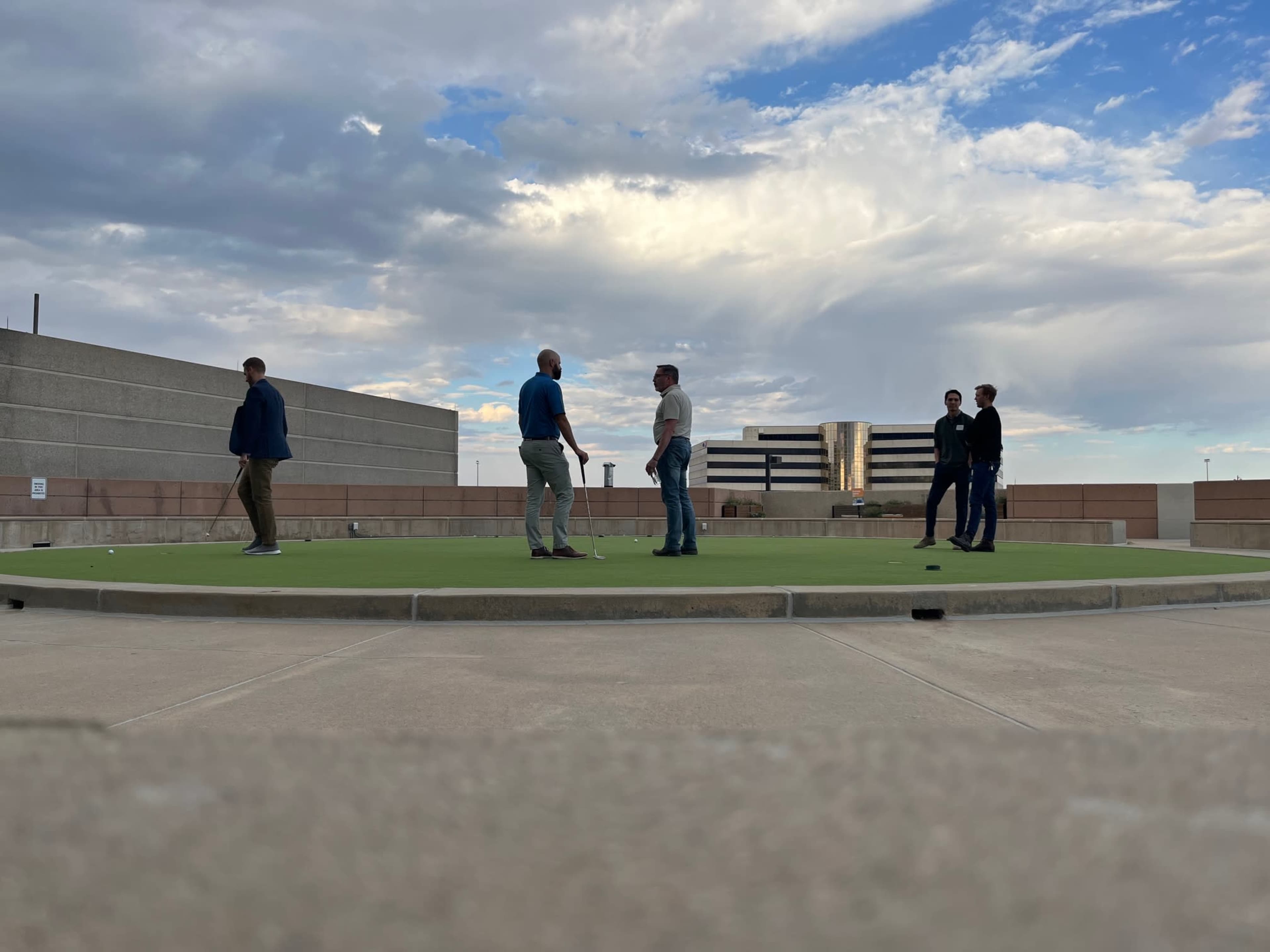 A group of five people practices putting on a rooftop golf green under a partly cloudy sky.