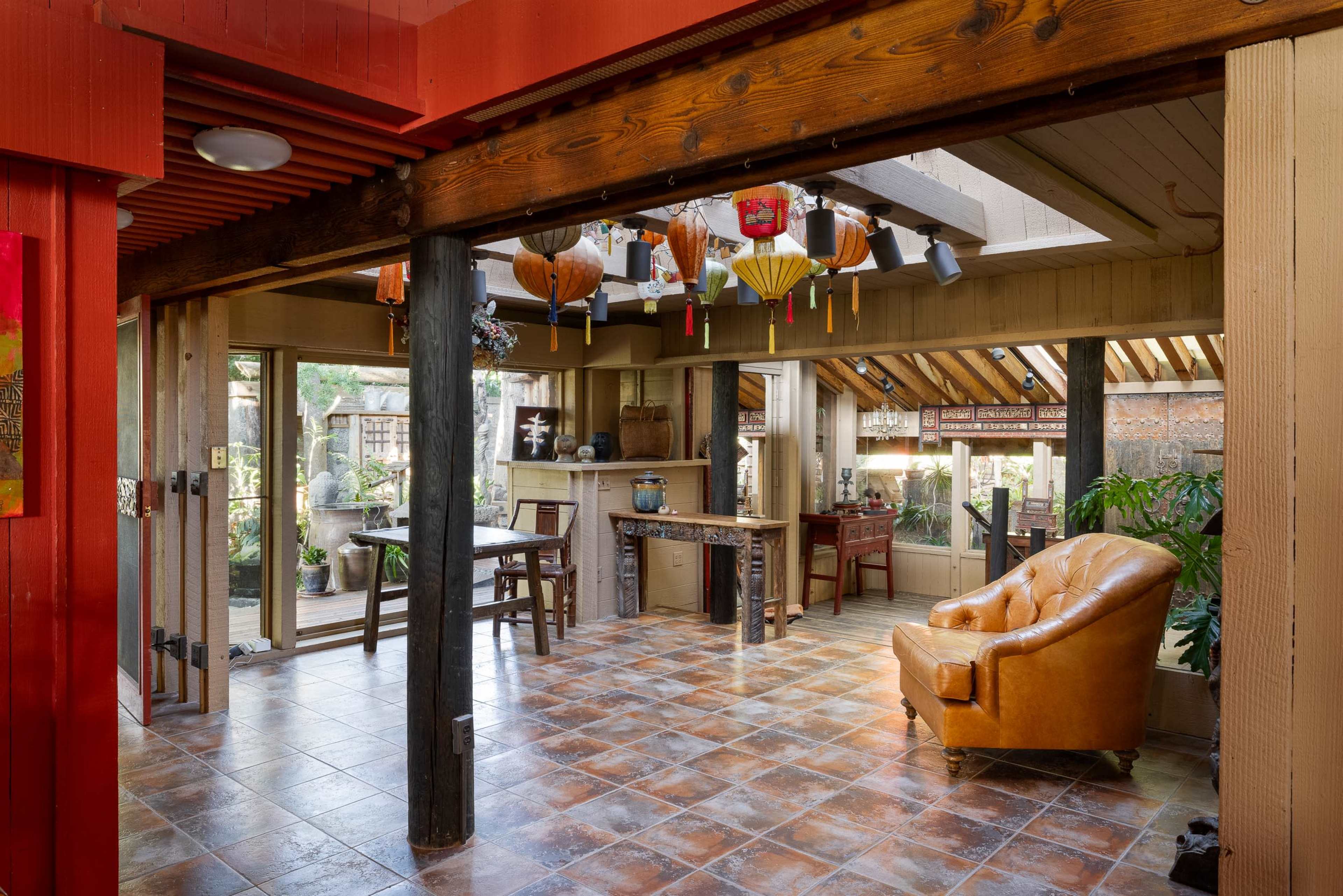 The image shows a spacious, open-concept living area featuring wooden beams, tile flooring, and a mix of traditional and contemporary furnishings, with large windows allowing natural light to enter.