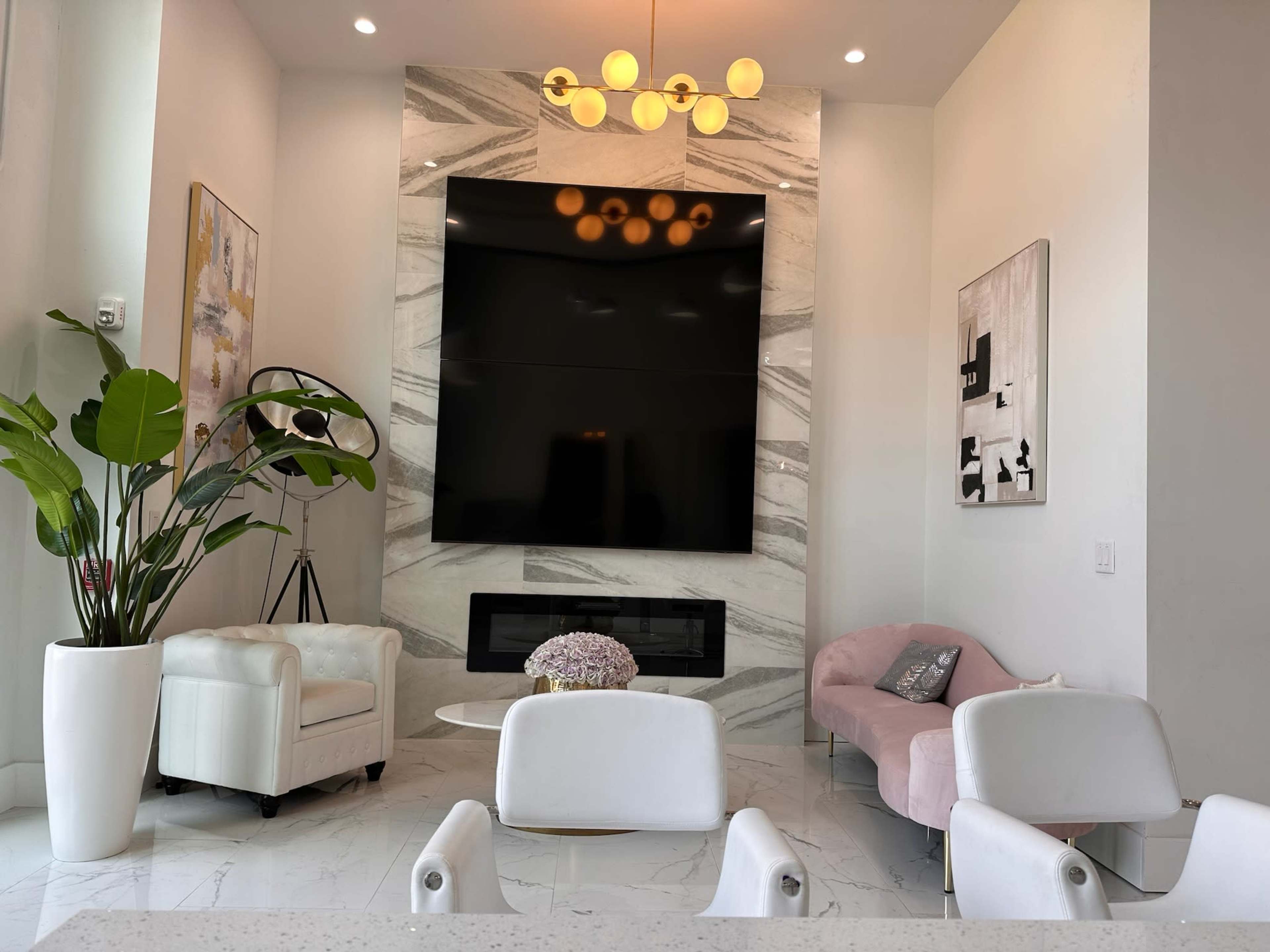 The image shows a modern living room with a large television mounted on a marble wall, two armchairs, a sofa, and a decorative plant in a white pot.